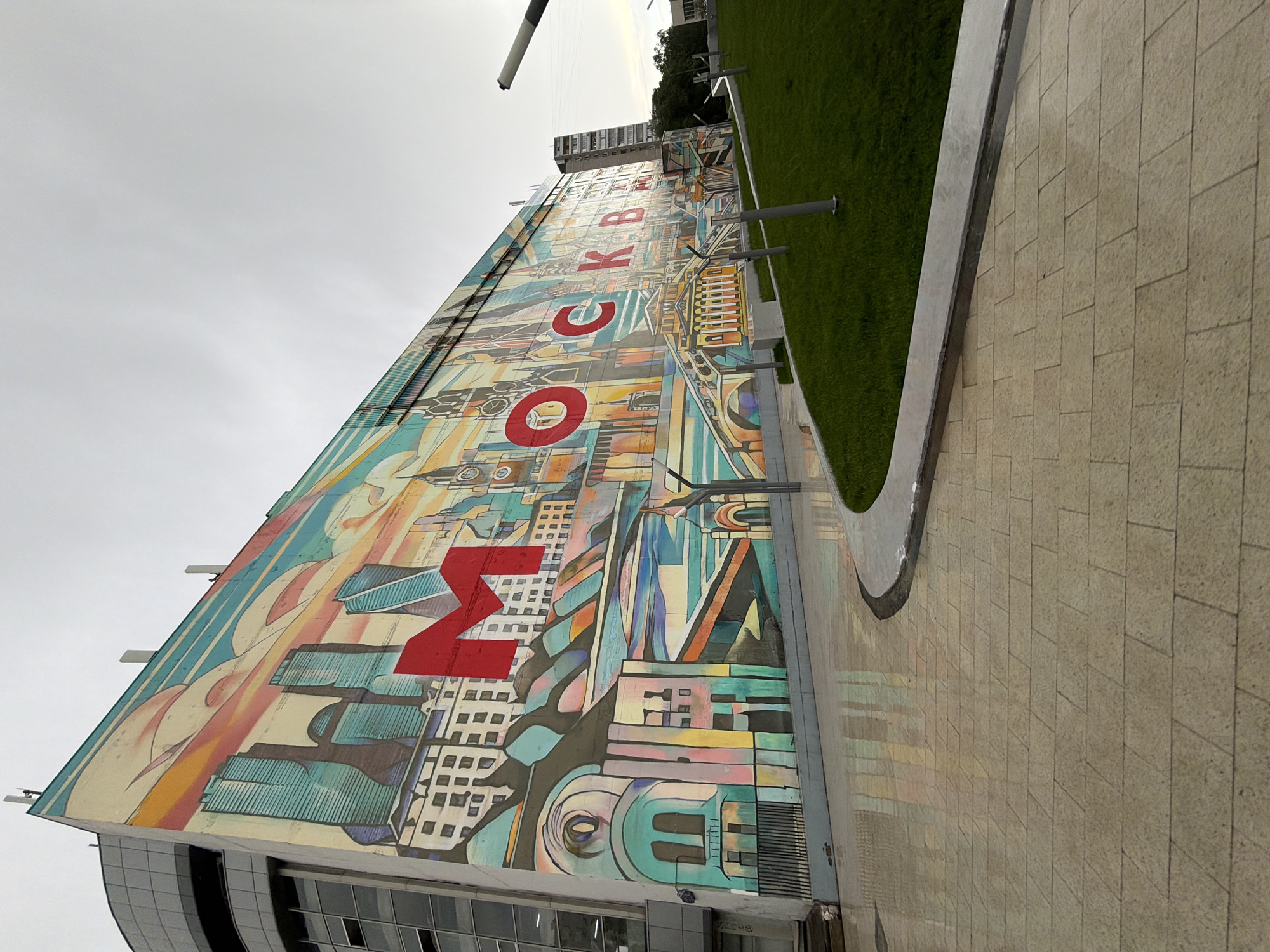 The image shows a building in Moscow, Russia, featuring a large mural. The mural incorporates the letters "M," "O," "K," and "B" in a stylized, artistic design. The cityscape elements within the mural suggest an urban theme. Adjacent to the building is a section of grass and a paved walkway. The overall scene is taken during daylight under a cloudy sky. The building's architecture is modern, with visible window structures.