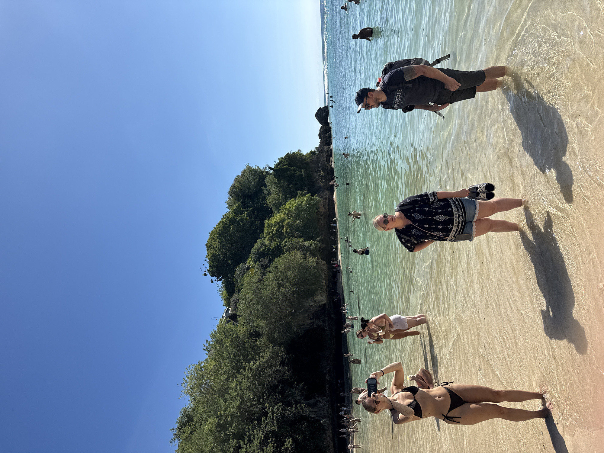 The image depicts a scene at Melasti Beach, located in Kuta Selatan, Indonesia. The clear turquoise water meets a sandy beach, with a lush green cliff face on the left side of the frame. Several people are wading and swimming in the water. In the foreground, a woman in a black bikini is holding up a smartphone to take a picture. Behind her, other beachgoers are visible. A man wearing a dark shirt, shorts, and a baseball cap is standing further into the water with a backpack. A woman with sunglasses, a black patterned shirt, and denim shorts stands next to the man, holding her shoes in her hand. The sky is clear and blue.