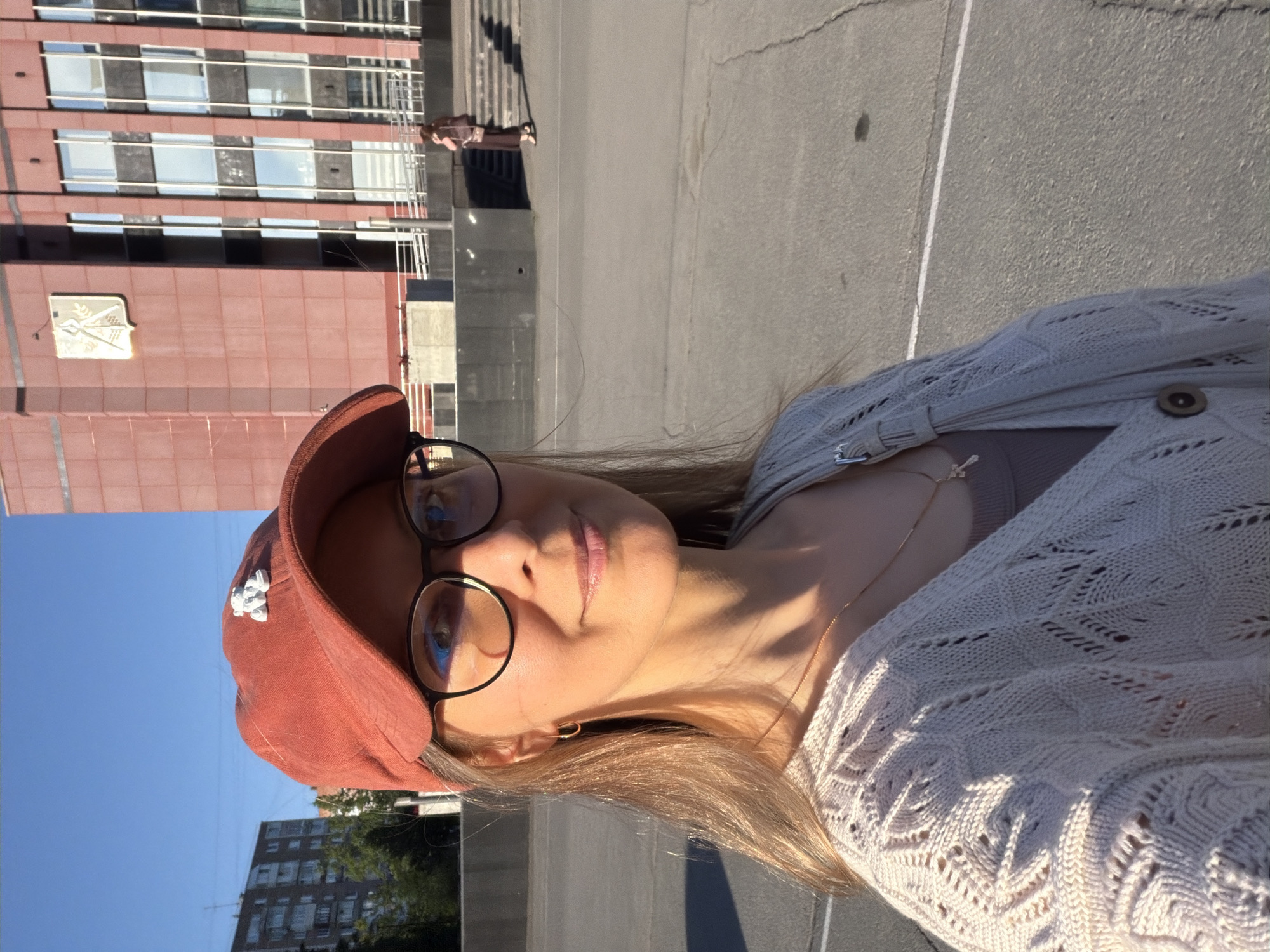 A woman with fair skin, wearing glasses and a rust-colored baseball cap, is the primary subject of the image. She is also wearing a light-colored, knitted cardigan. The background features a building with a rust-colored facade and numerous windows, a section of concrete pavement, and a glimpse of a blue sky. An emblem is visible on the building's facade. The overall setting appears to be in Izhevsk, Russia, given the building's architectural style and the overall aesthetic of the surroundings. A person is walking up a set of stairs in the background.