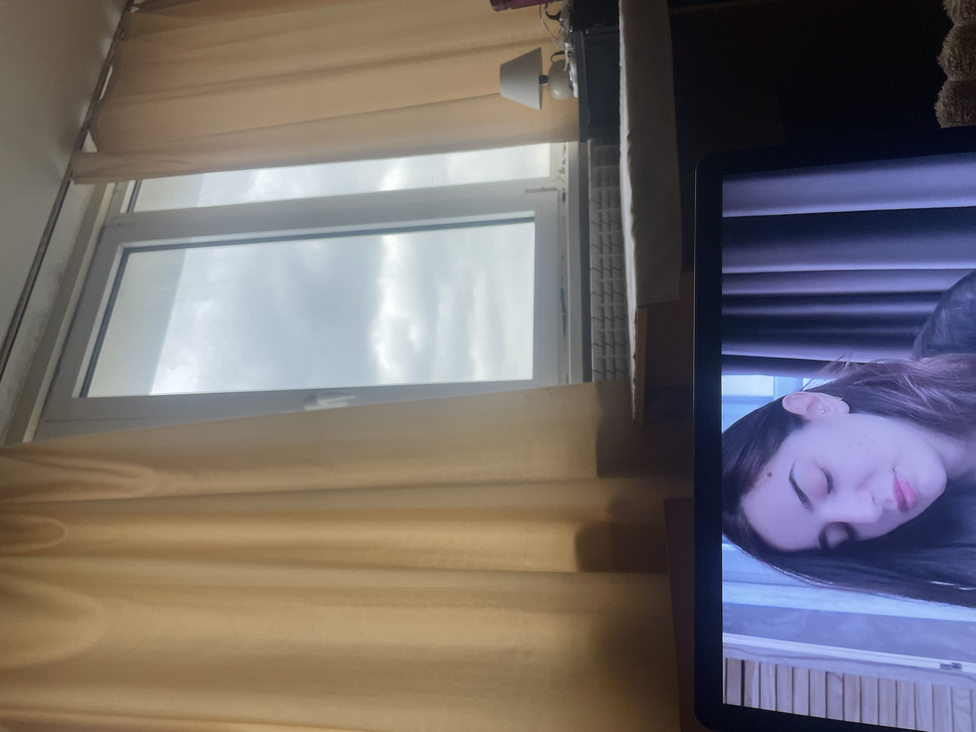 The image shows a window with light beige curtains partially obscuring the view outside. The window reveals a cloudy sky. To the right of the window, there is a screen displaying a person's face. The scene appears to be indoors, possibly a room in an apartment or house. It is not possible to determine a specific location in Varna, Bulgaria from this image.