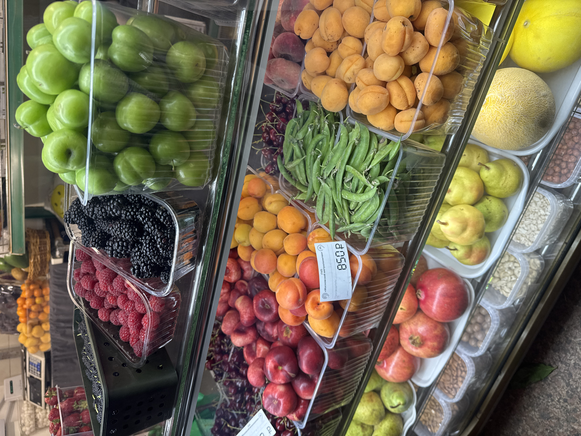 The image shows a fruit and vegetable display at a store in Moscow, Russia. The items are neatly arranged in clear plastic containers on a tiered shelf system. Fruits visible include green plums, blackberries, raspberries, strawberries, apricots, peaches, cherries, nectarines, pomegranates, pears, and a melon. Green peas are also displayed. A price tag is visible for one of the apricot varieties, indicating the cost is 850 rubles. The display is indoors, likely within a market or grocery store. The overall scene conveys a well-stocked and organized retail environment.