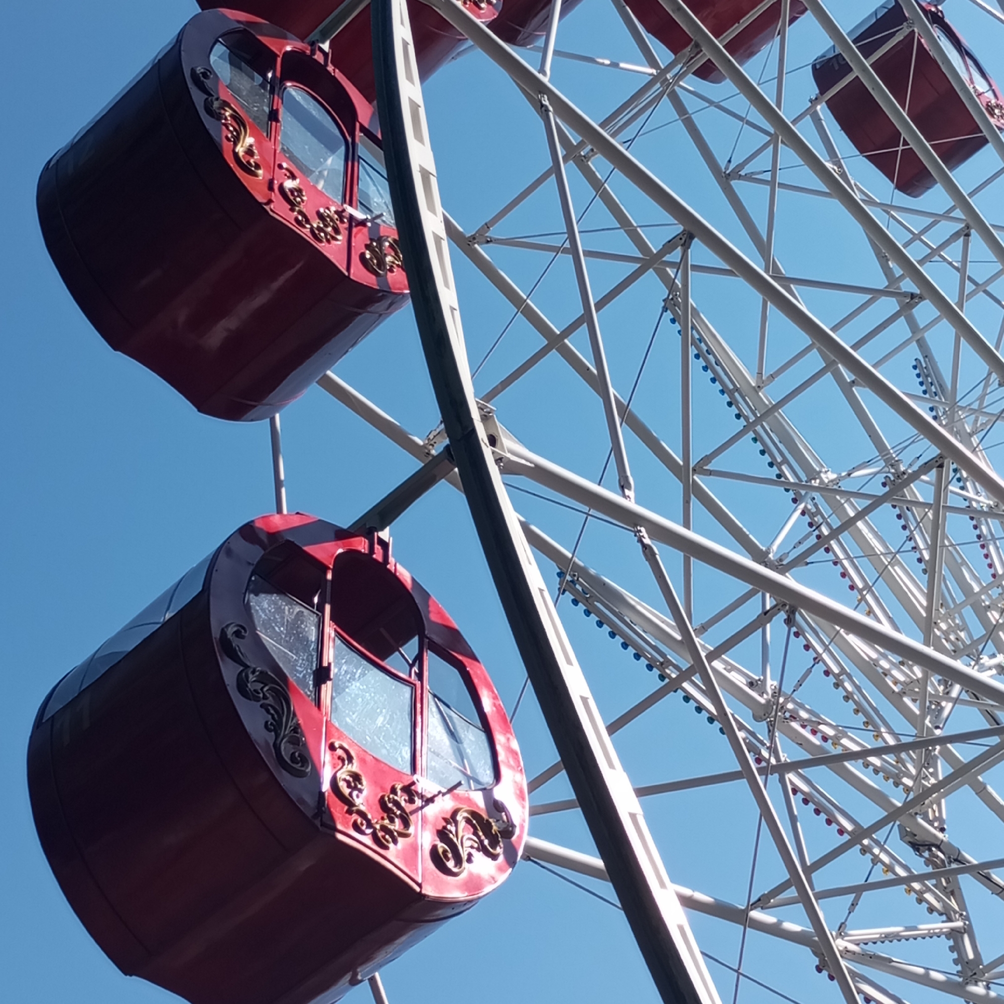 The image shows two red gondolas of a Ferris wheel against a clear blue sky. The Ferris wheel structure is white and made of metal beams. The gondolas are oval-shaped with glass windows and ornate gold detailing. One gondola is positioned higher and to the left of the frame, while the other is lower and to the left. The background is completely the sky, with no other elements visible. The image is taken from a low angle, looking up at the Ferris wheel. This Ferris wheel is located in Izhevsk, Russia.