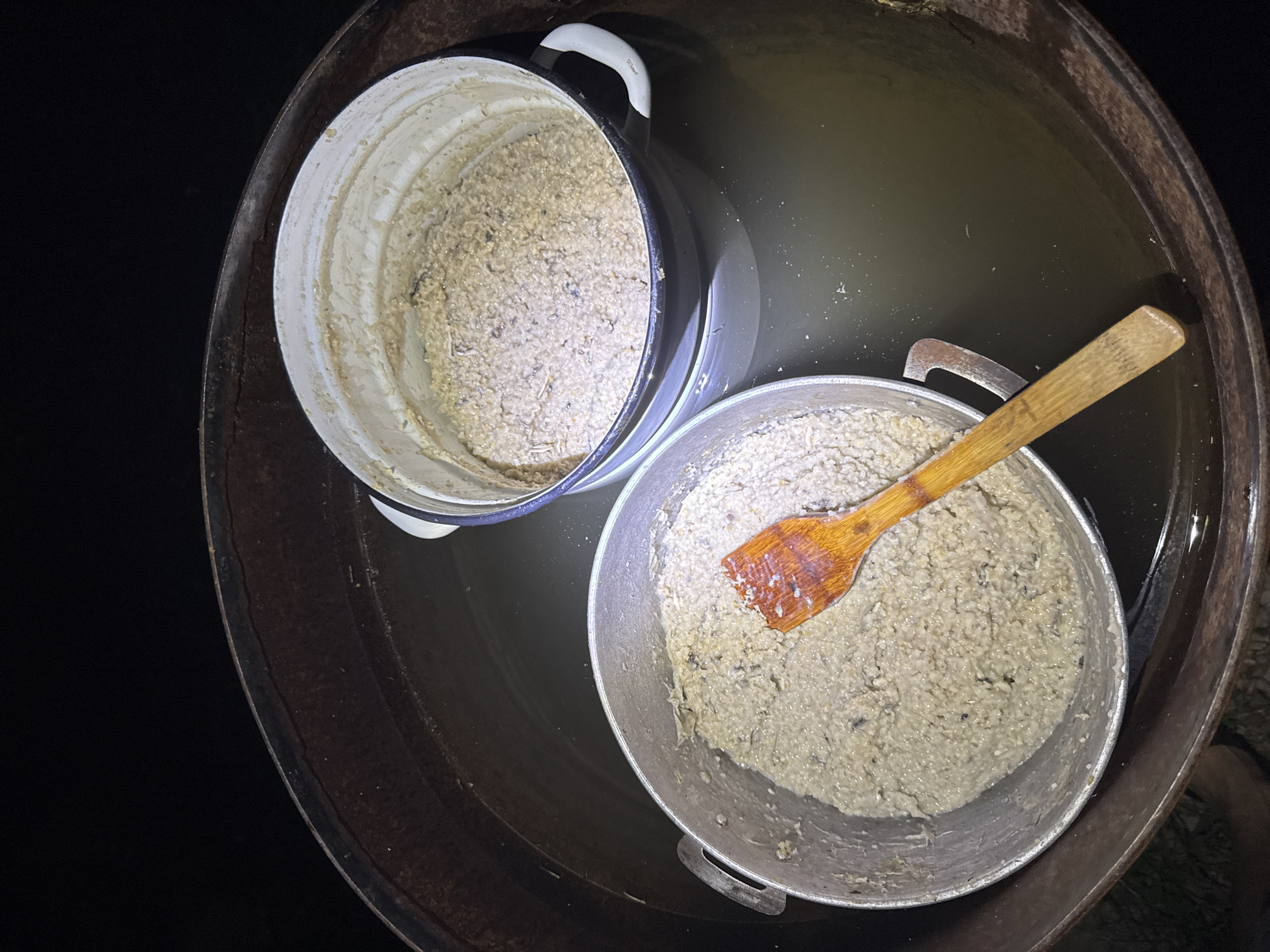The image shows two metal pots filled with a light-colored, textured food substance, possibly porridge or a grain-based mixture. One pot, made of silver-colored metal with handles, is in the foreground on the left, and a wooden spatula is resting inside it. Behind and slightly to the right is a white enamel pot with black trim and handles, also filled with the same food substance. Both pots are situated inside a large, dark-colored, round container, possibly a barrel, which is partially filled with murky water. The background is dark, suggesting the scene is outdoors at night or in a dimly lit area. The location is identified as Dobrovske rural settlement, Ukraine.