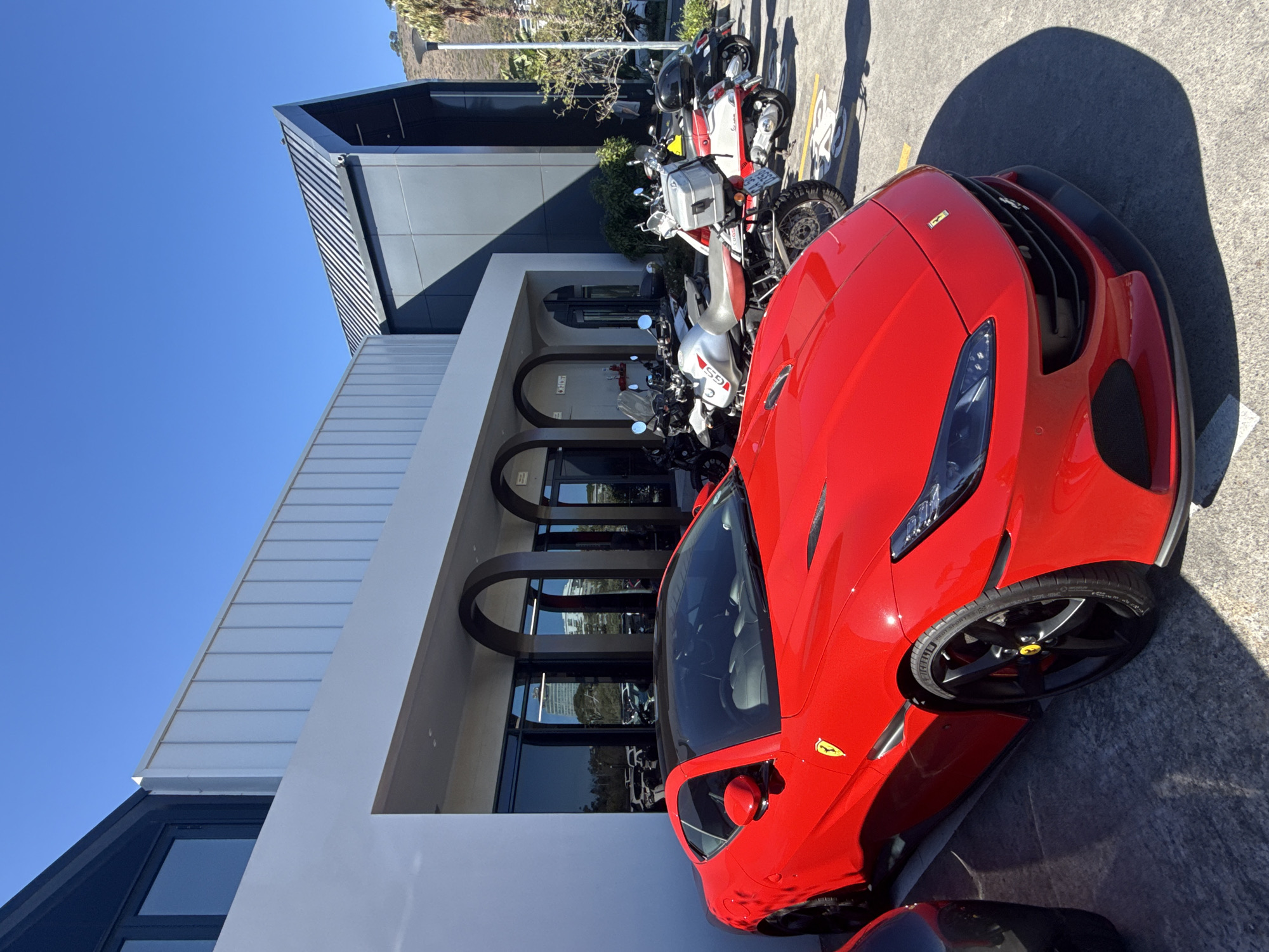 A red Ferrari is parked on concrete outside a modern building with a combination of grey, white, and dark blue/black cladding under a clear blue sky. Behind the car are several motorcycles parked in a line. The architecture suggests a contemporary design with arched window openings. The image appears to have been taken in an urban setting. The location in Cape Town, South Africa cannot be verified based solely on the image.
