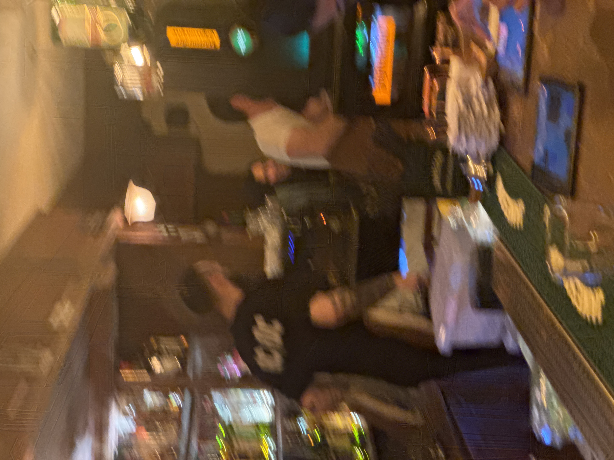 The image shows the inside of a bar, possibly in Yerevan, Armenia, based on the request. A man in a black t-shirt with "AC/DC" printed on it is standing at the bar. Another person in a white shirt is also present at the bar. Bottles of alcohol are visible on the bar top, and there are screens behind the bar. The lighting is dim.