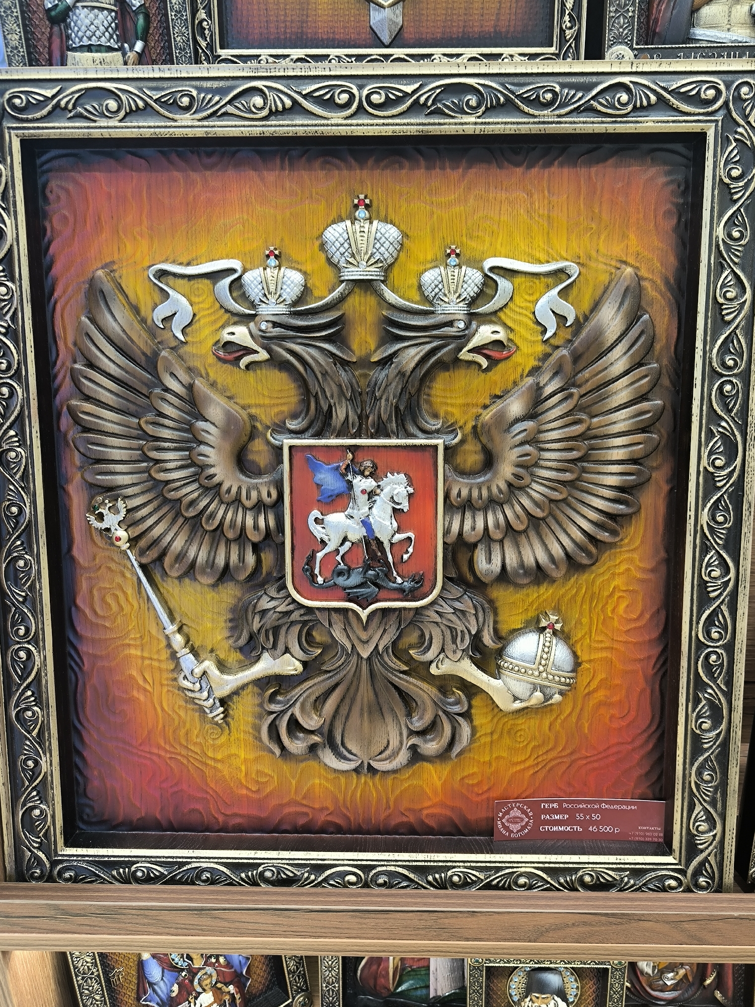 The image shows a close-up of a carved wooden relief of the Coat of Arms of the Russian Federation. The emblem features a double-headed eagle with two crowns, each adorned with a cross. The eagle's wings are spread wide, and in its talons, it holds a scepter on the left and an orb on the right. A shield is superimposed on the eagle's chest, depicting Saint George slaying a dragon. The background of the relief is a gradient of warm colors, transitioning from yellow at the top to red and orange at the bottom, with carved wood grain visible. The entire relief is framed by an ornate, dark-colored border with gold accents. A small label is affixed to the bottom right of the relief, bearing text in Russian.
While the image itself does not provide specific location information within Novosibirsk, Russia, it is a common type of souvenir or decorative item found in souvenir shops or craft markets in tourist areas. The label text, partially legible, indicates it is the "Coat of Arms of the Russian Federation" with dimensions "55x50" and a "cost" of "46,500 p." (likely rubles).