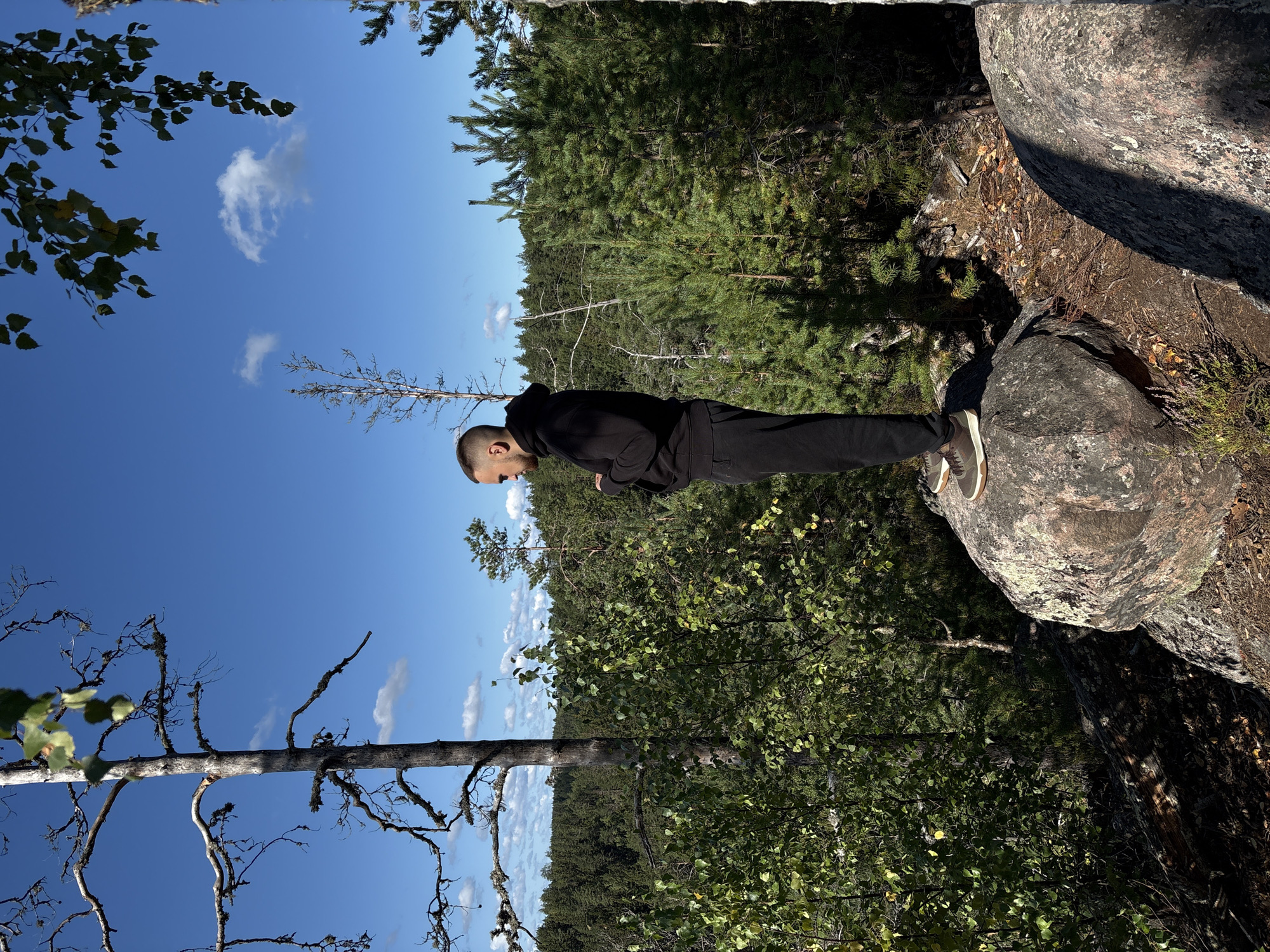 A young man wearing a black hoodie and black pants stands on a large rock in a forest. He is looking down at his phone. The forest is dense with pine trees and some deciduous trees. The sky is blue with some white clouds. The man is positioned on the right side of the frame, and the forest stretches out behind him. The image appears to have been taken in the city of Kir'yavalakhti, Russia.