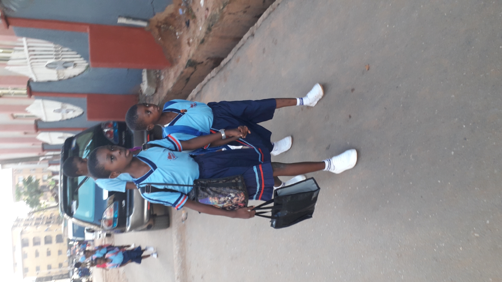 A group of children in school uniforms walks along a city street in Onitsha, Nigeria. They wear blue and dark blue uniforms with white socks and white shoes. Some hold hands and carry bags. The setting includes buildings, cars, and a paved street typical of an urban environment.