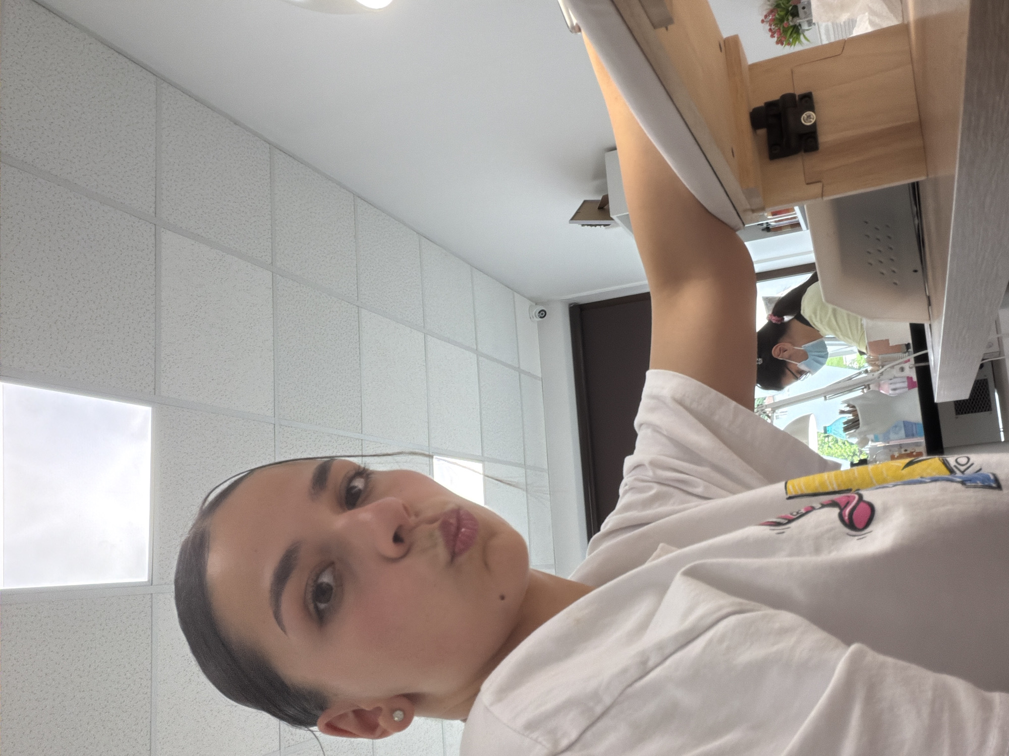 The image shows a young woman with dark hair pulled back, wearing a white t-shirt with a colorful graphic, posing with her lips pursed. She is seated at a table in what appears to be a nail salon. In the background, another woman, wearing a mask and glasses, is working at a manicure station. The environment includes a tiled ceiling with fluorescent lighting, a security camera mounted on the wall, and various manicure supplies, including bottles, tools, and a nail lamp. A wooden armrest is positioned in the foreground. The image does not provide any specific information to indicate it is located in the city of Gabrovo, Bulgaria.
