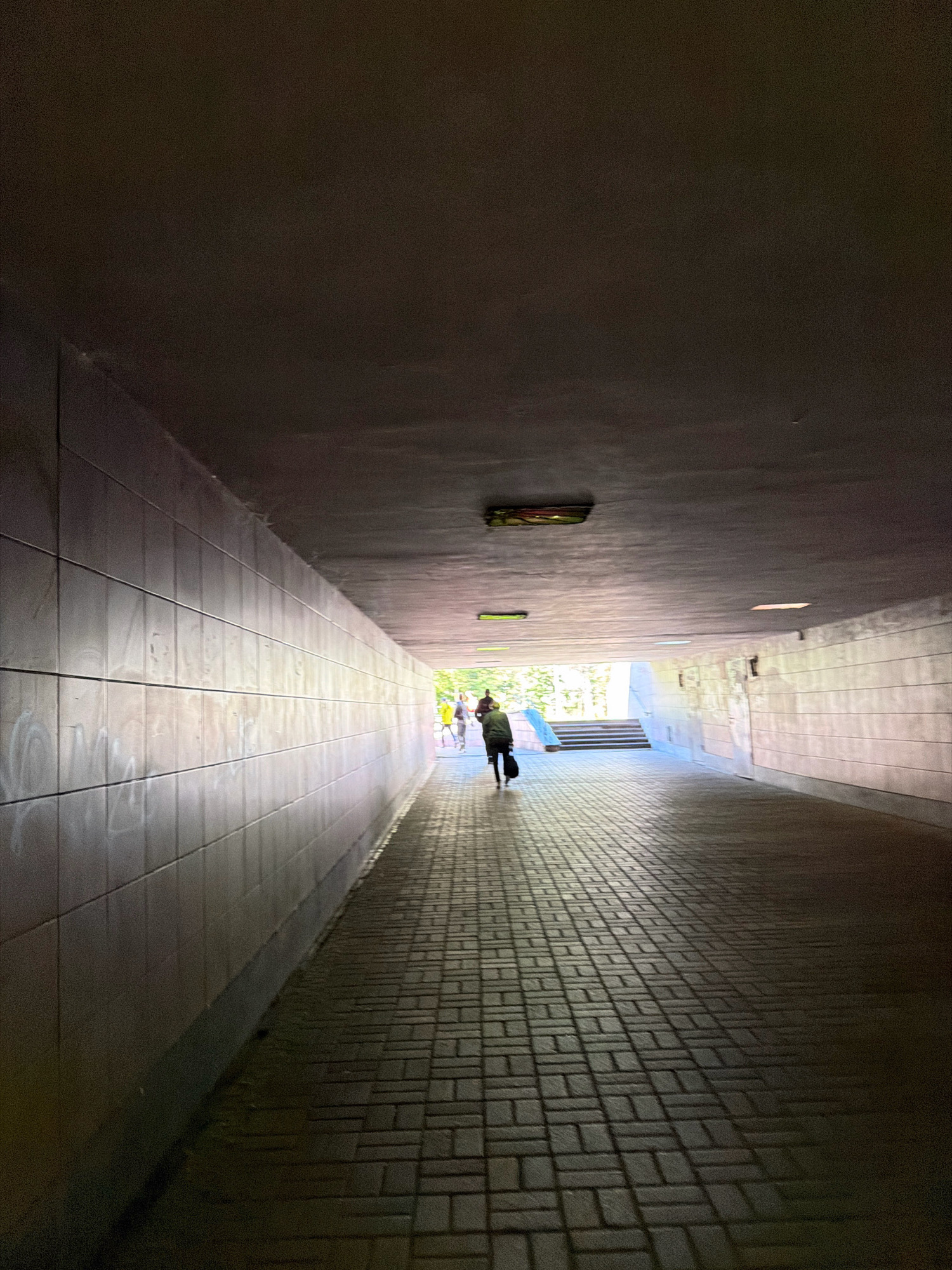 This image captures an underground pedestrian passage, commonly known as a underpass, in Kyiv, Ukraine. The passage is constructed with concrete walls tiled with rectangular, light-colored tiles, some of which show faint graffiti. The floor is paved with rectangular bricks laid in a herringbone pattern. The ceiling of the underpass is dark and unadorned, with two visible rectangular light fixtures, one on the left side and another on the right, along with a larger, elongated fixture on the ceiling further ahead, illuminated with a greenish hue.
In the distance, at the end of the underpass, daylight illuminates the scene. A person is seen walking away from the camera towards the light. This individual is wearing dark clothing and carrying a dark bag over their shoulder. Further ahead, other figures are visible, some in bright yellow clothing, suggesting they are also moving towards the exit. Stairs are visible to the right of the distant figures, leading upwards and out of the underpass. The lighting within the underpass creates a strong contrast between the dimly lit interior and the bright exterior. The overall impression is that of a functional urban passage connecting two points of the city.