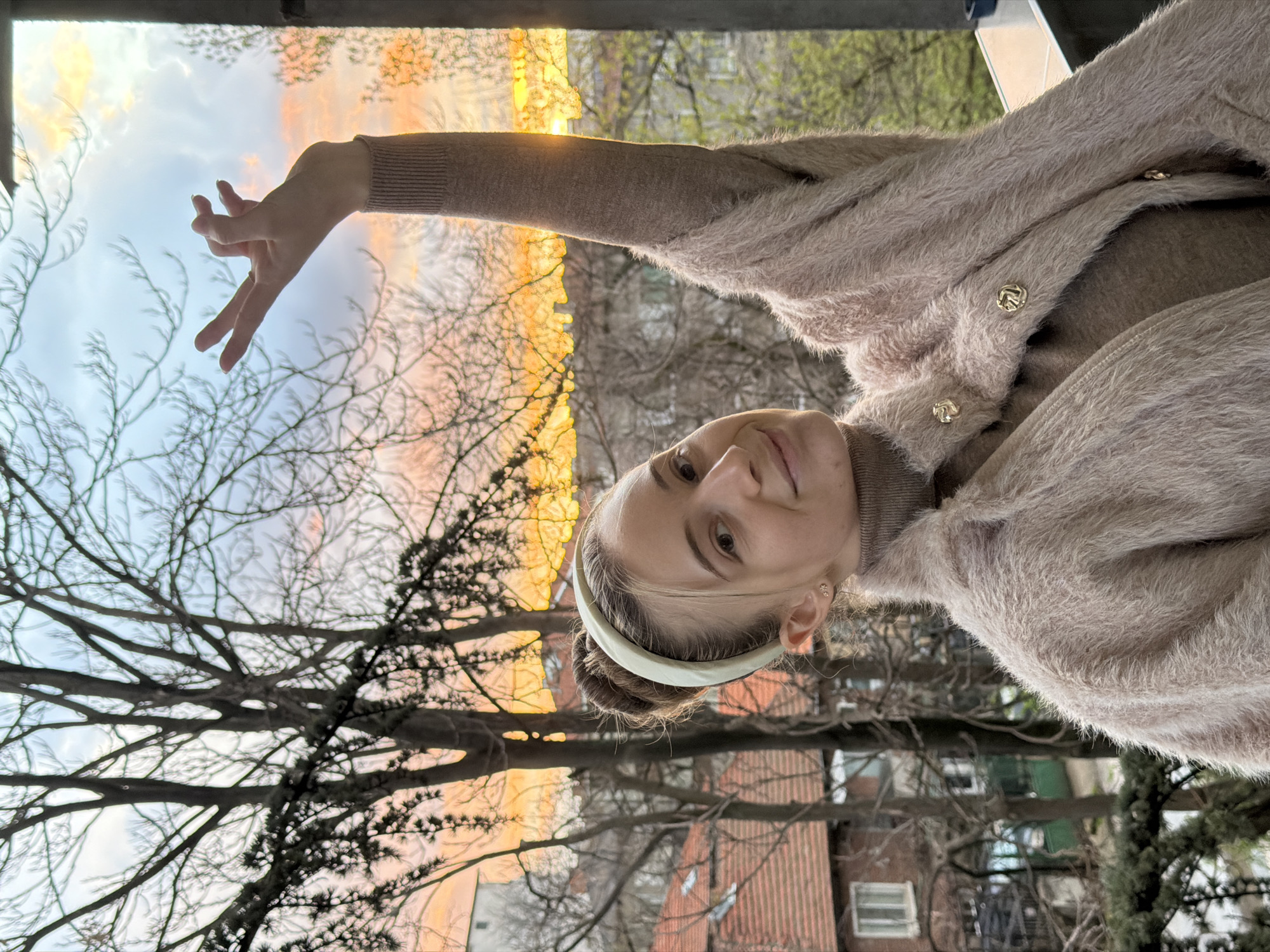 The image shows a woman with light skin and hair pulled back with a white headband, wearing a brown turtleneck sweater and a fluffy light brown cardigan with gold buttons. She is positioned outdoors with bare tree branches visible behind her. Her right arm is raised towards a brightly lit sky with streaks of orange and yellow. In the background, there are buildings with red roofs and windows. The location is likely an urban setting in Plovdiv, Bulgaria, based on the architectural style of the buildings.