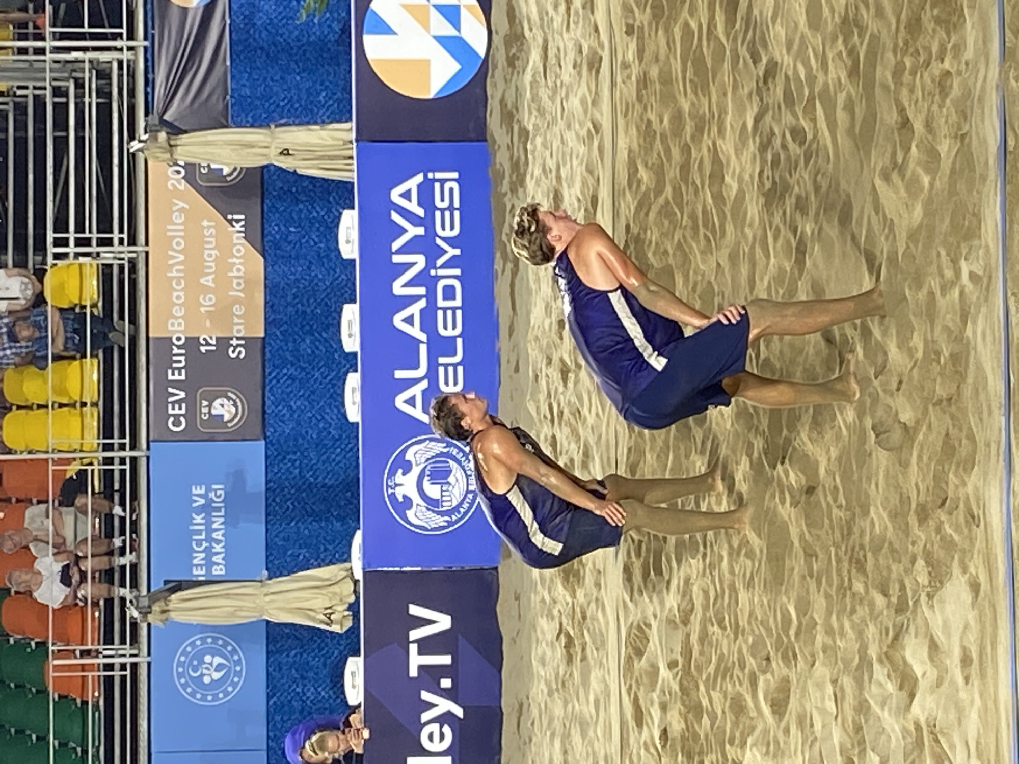 This image depicts a beach volleyball match in Alanya, Turkey. Two male athletes, both fair-skinned and visibly sweating, are positioned on a light brown sand court, bent forward with hands on their knees, in a ready or resting stance. The athlete on the left has dark hair, wearing a dark blue and white tank top and dark shorts, and is looking towards the right side of the court. The athlete on the right has lighter, curly hair, also in a dark blue and white tank top and dark shorts, and is looking towards the left.
In the background, tiered stadium seating with green, orange, and yellow plastic chairs is visible, occupied by several spectators. Notably, two older individuals are seated in orange chairs in the upper left, and a young child with blonde hair is engrossed with a device behind a banner in the lower left.
Prominent advertising banners line the upper and lower sections of the background. The upper banners display "GENÇLİK VE BAKANLIĞI" (Ministry of Youth and Sports) with an accompanying logo, and "CEV EuroBeachVolley 20..." with "12 - 16 August Stare Jablonki" (likely an advertisement for a different event). The lower banners feature "...ley.TV", "T.C. ALANYA BELEDİYESİ" (Republic of Turkey, Alanya Municipality) with a distinct castle/fortress logo, and a colorful geometric logo. A collapsed, beige umbrella is secured to a pole, positioned vertically between the banners. The scene is brightly and evenly lit, suggesting artificial lighting for an evening game or indoor venue.