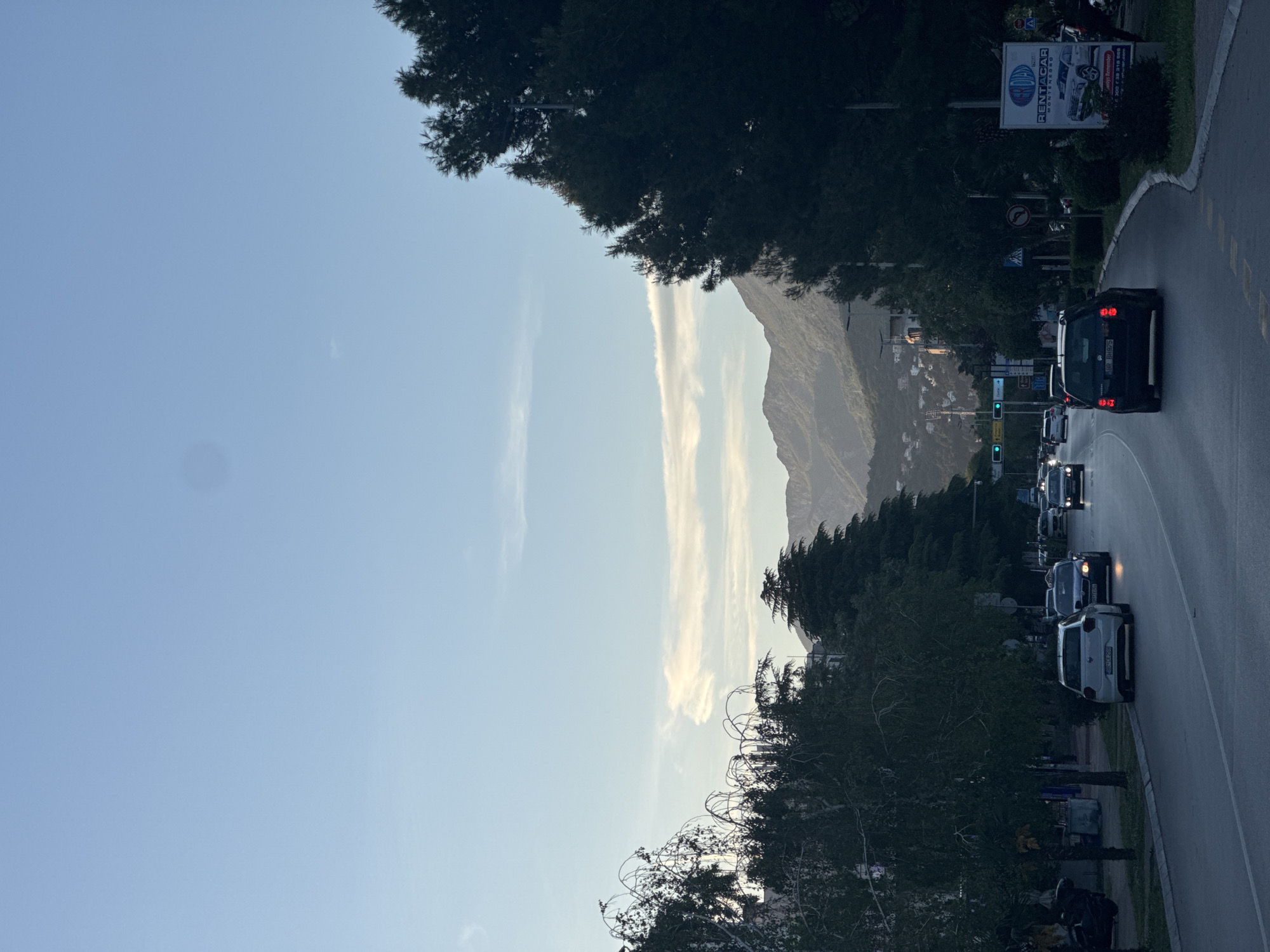 The image, rotated counter-clockwise, depicts a scenic view of a road winding through a coastal or mountainous region, likely in Bar, Montenegro, as indicated by visible signage. The scene captures a late afternoon or early evening ambiance, characterized by soft, clear blue skies with elongated white clouds hovering above distant mountains.
In the foreground, a paved road curves gently to the right, with several vehicles visible. A white SUV is parked or moving slowly on the left side of the road, followed by a dark sedan. Further along, a black SUV with illuminated taillights appears to be stopped at a traffic light, which displays a green signal. Other cars are visible in the distance. The road itself features dashed white lines on its right edge.
The roadside is lined with lush, dark-green trees and foliage, providing a sense of natural beauty. On the right side of the road, a large billboard stands prominently, displaying "RENTACAR MONTENEGRO" with images of cars, and the word "TAXI" also visible. Several road signs are also present, including a "No Entry" and a "No Left Turn" sign.
In the background, majestic mountains rise under the clear sky, with scattered buildings and structures dotting their lower slopes, suggesting human habitation integrated into the natural landscape. The overall impression is one of a peaceful, ordinary day in a picturesque setting with routine traffic flow.