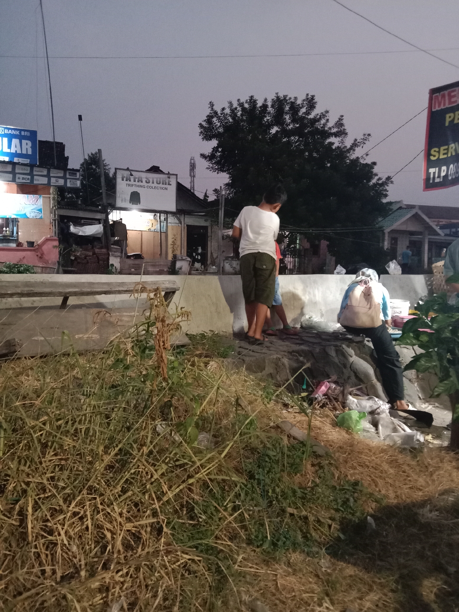 In Langensari, Indonesia, at dusk, a group of people is gathered near a concrete embankment. A young person in a white t-shirt and dark shorts stands facing away from the camera, looking towards a building with a sign that reads "FATA STORE TRIFTING COLECTION". Another person, wearing a light blue shirt and a light brown backpack, is partially obscured by the embankment, facing the opposite direction. They appear to be rummaging through a pile of trash. Nearby, a younger child in a white shirt and red shorts is also present. The ground is covered with dry grass and debris, including several plastic bags and other discarded items. In the background, several businesses are visible, including one with a "BANK BRI" sign. The sky is overcast, indicating the end of the day.