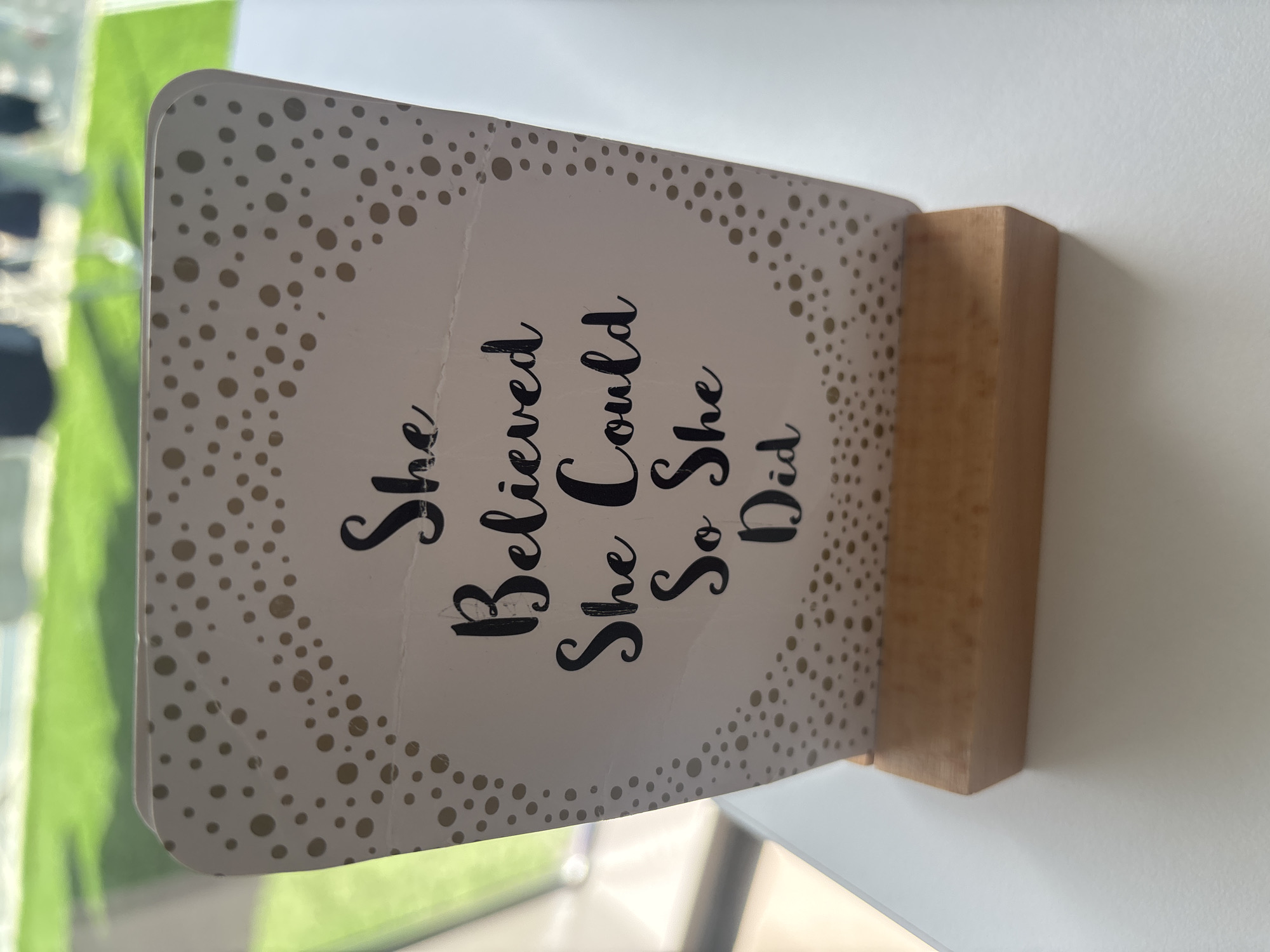 A white card with gold polka dots and the quote "She Believed She Could So She Did" is standing upright in a wooden base. The card is positioned on a white table. In the background, out of focus, is a green lawn and blurred figures of people, suggesting an outdoor setting. There is no indication of the city of Dubai or the United Arab Emirates in the image.