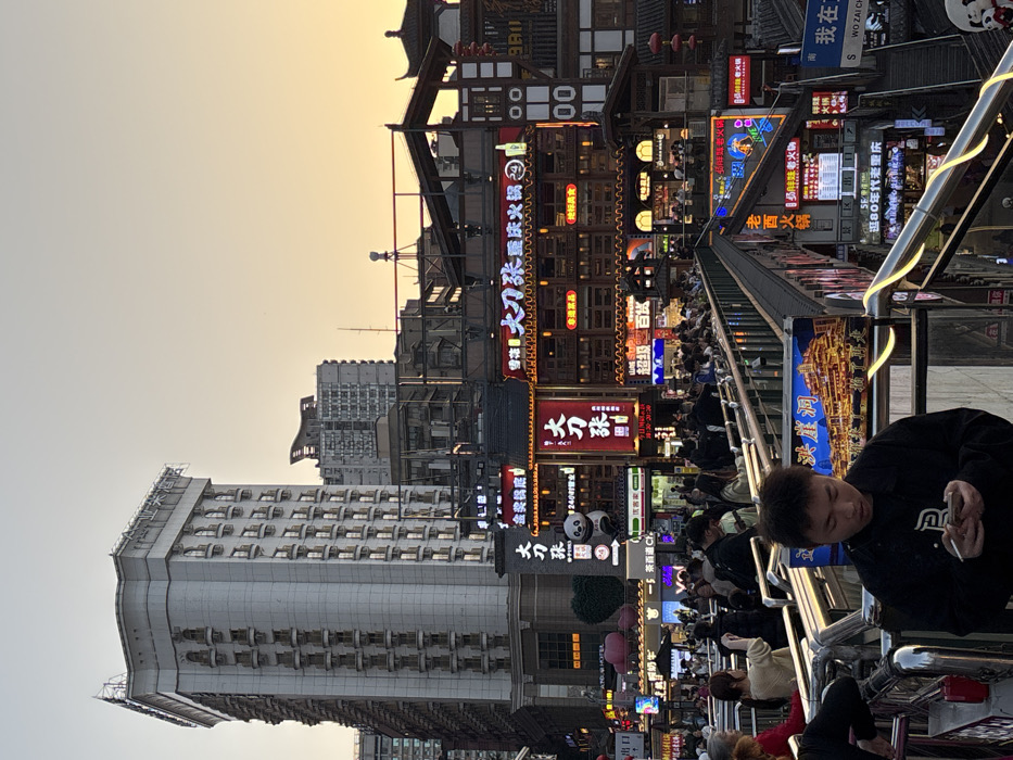 The image captures a vibrant urban scene at dusk in Hongyadong, Yuzhong District, Chongqing, China. A tall, light-colored modern skyscraper with numerous small, arched windows dominates the left background, its top featuring a decorative crown and rooftop signage, set against a clear sky exhibiting a gradient from pale blue to orange-yellow. Below and to its right, a multi-tiered complex of traditional Chinese-style wooden buildings, characteristic of 'diaojiaolou,' stands extensively illuminated by numerous red and yellow neon signs. These signs display Chinese characters, including "大刀张重庆火锅" (Big Knife Zhang Chongqing Hotpot) and "洪崖洞" (Hongyadong), with a prominent panda mascot visible near a shop entrance. Red lanterns hang from the traditional structures on the far right.
An elevated pedestrian walkway with polished metal railings featuring integrated yellow LED strip lighting stretches horizontally across the mid-ground. This walkway is densely populated with an indistinct crowd of casually dressed individuals, either walking or standing, observing the surroundings.
In the immediate foreground, a young male, approximately late teens to early twenties, is centered, wearing a dark jacket. He looks down, engrossed with a smartphone held in both hands, with a lit cigarette visible between the fingers of his right hand. He stands next to the illuminated railing, which also displays a large signboard featuring an image of the illuminated Hongyadong complex. Artificial lights from businesses and signs extensively illuminate the scene.