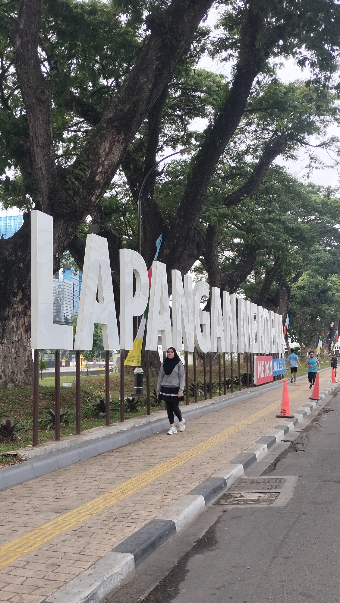 In City of Medan, Indonesia, at Lapangan Merdeka, a person in a grey sweater and black skirt with a black hijab walks on a brick sidewalk. The large white letters spelling "LAPANGAN MERDEKA" stand behind her, with a street and several orange traffic cones visible to the right. The background features large trees with lush green foliage and a building. The sky is overcast.