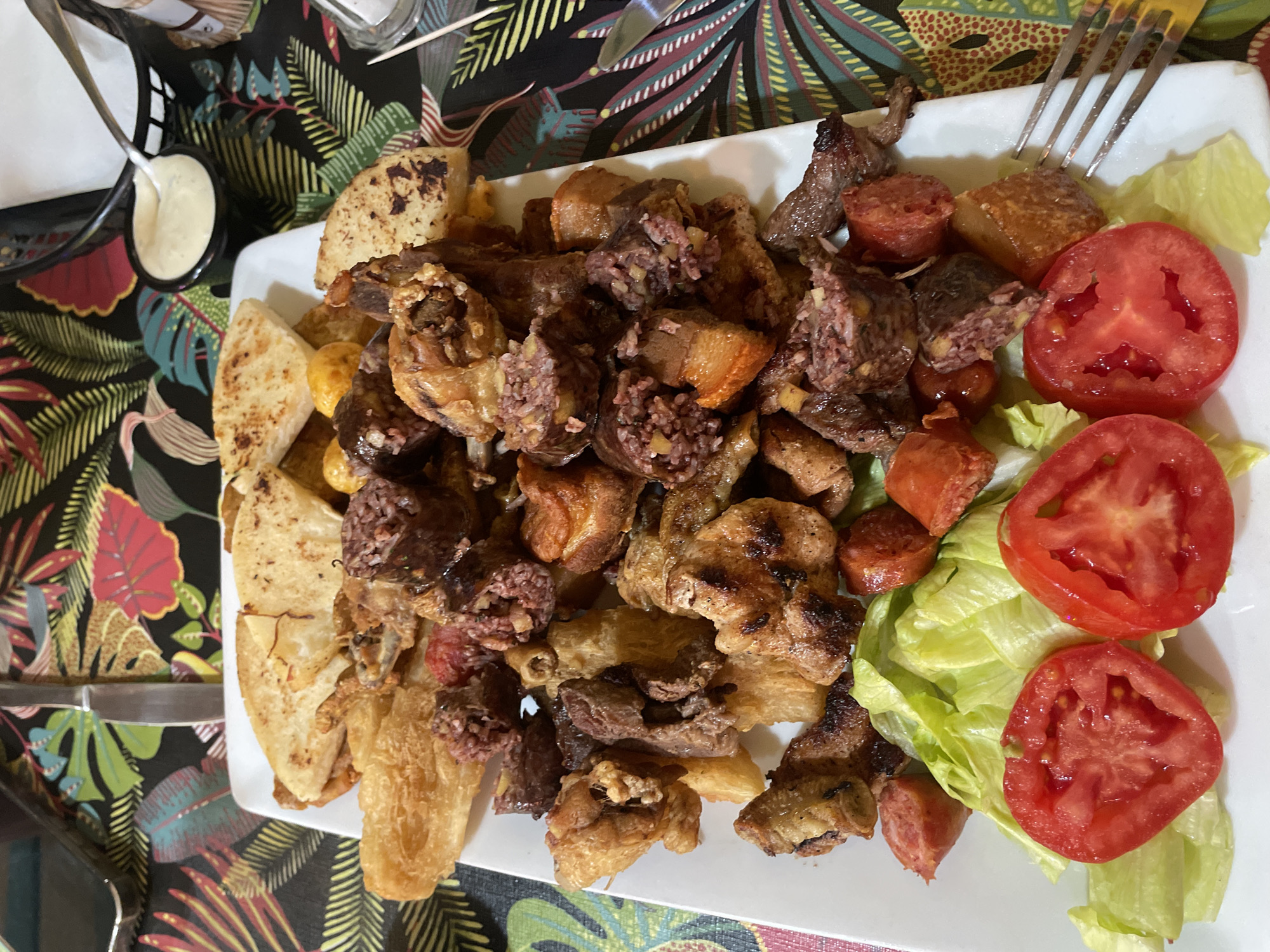 A white rectangular plate sits on a table with a dark floral tablecloth. The plate contains an assortment of cooked meats, sausage slices, fried bread slices, and three tomato slices on a bed of lettuce. A small container of white sauce with a spoon rests to the left of the plate. A fork is visible at the upper right corner of the plate. The scene is likely a meal setting in L'Hospitalet de Llobregat, Spain. There is no additional information to confirm this location, however. No people are visible.