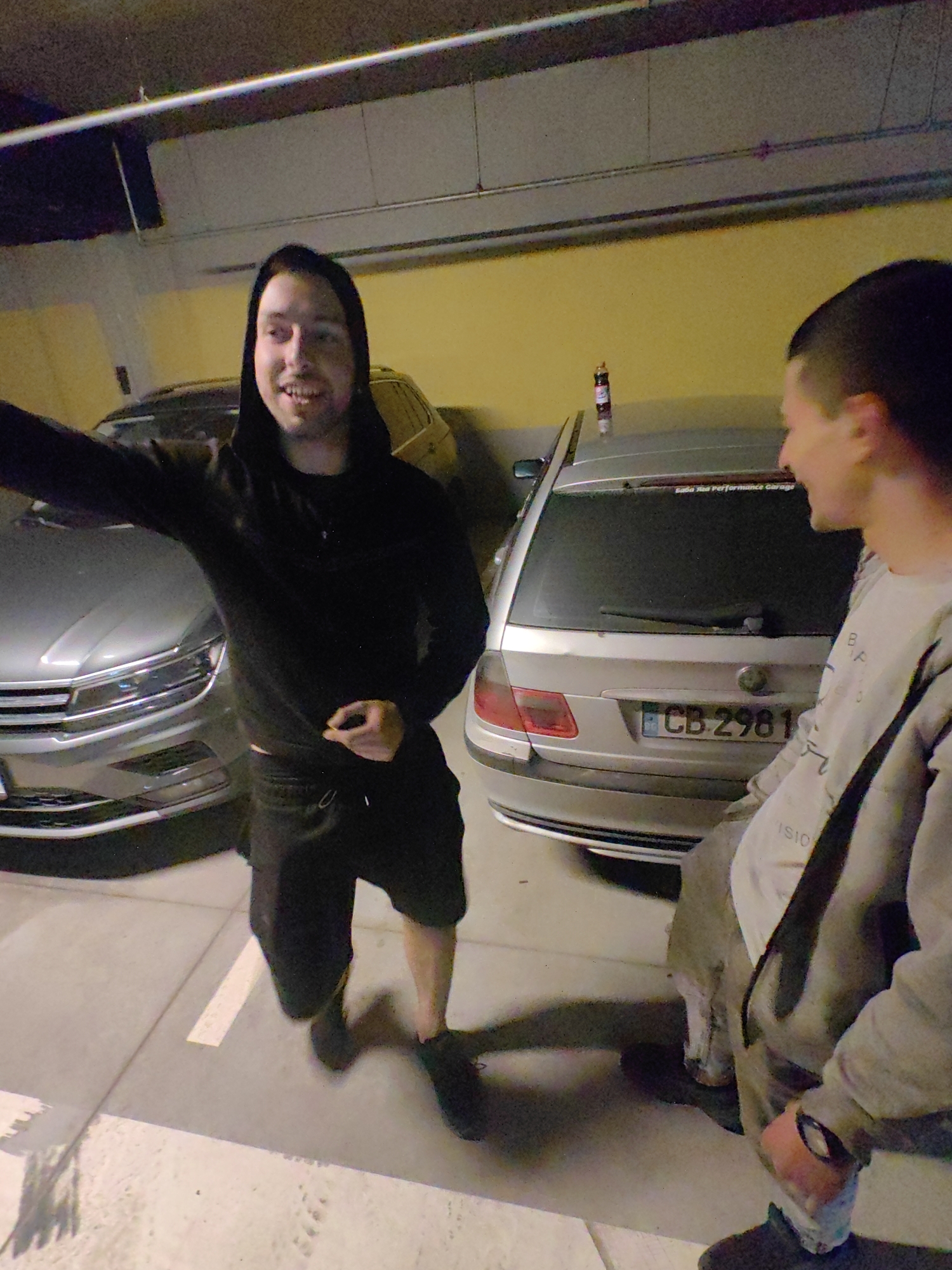 The image depicts two young men in an indoor parking garage, likely in Sofia, Bulgaria, as evidenced by the car's license plate "CB 2981," which is a Sofia license plate. The man on the left wears a black hooded sweatshirt and black shorts. He is gesturing with his left arm. The man on the right is wearing a light-colored sweatshirt and has short hair. A silver car and a black SUV are visible in the background, parked in adjacent parking spaces. There is a brown bottle on the trunk of the silver car.