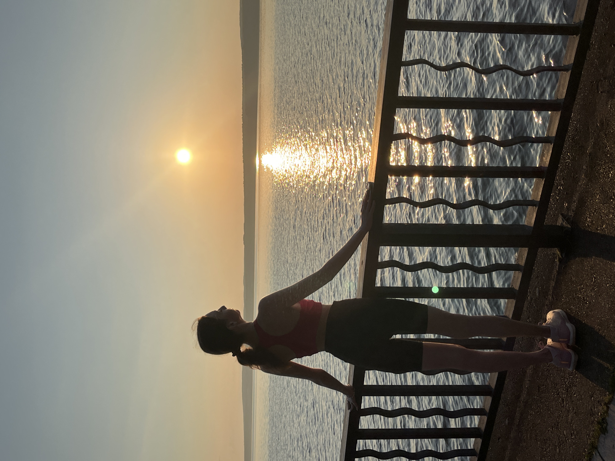 A young woman with a ponytail, wearing a red sports bra and black athletic shorts, stands at a dark metal railing overlooking a body of water. Her back is to the camera as she gazes at the sun setting over the horizon, casting a shimmering reflection on the water. The sky is a gradient of pale blue and soft orange. The railing has decorative, wavy vertical bars. The ground beneath her appears to be a textured surface, possibly asphalt or concrete, with some grass or weeds at the edge. The scene suggests an outdoor recreational area, likely a promenade or embankment. This location is the embankment of the Kama River in Chaikovsky, Russia.