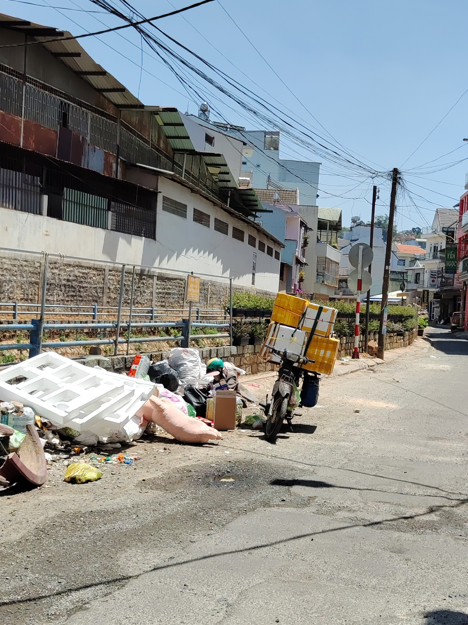 The image depicts a street scene in Da Lat, Vietnam. A pile of garbage is on the side of the street, including styrofoam, plastic bags, and other discarded items. A motorcycle laden with several yellow boxes is parked nearby. Overhead, a dense network of electrical wires crosses the sky. The buildings lining the street are a mix of architectural styles. The street surface is asphalt and appears worn. A red and white striped pole is visible further down the street. A few people are seen in the distance on the right side of the street. A railing separates the street from a waterway. The weather appears sunny.