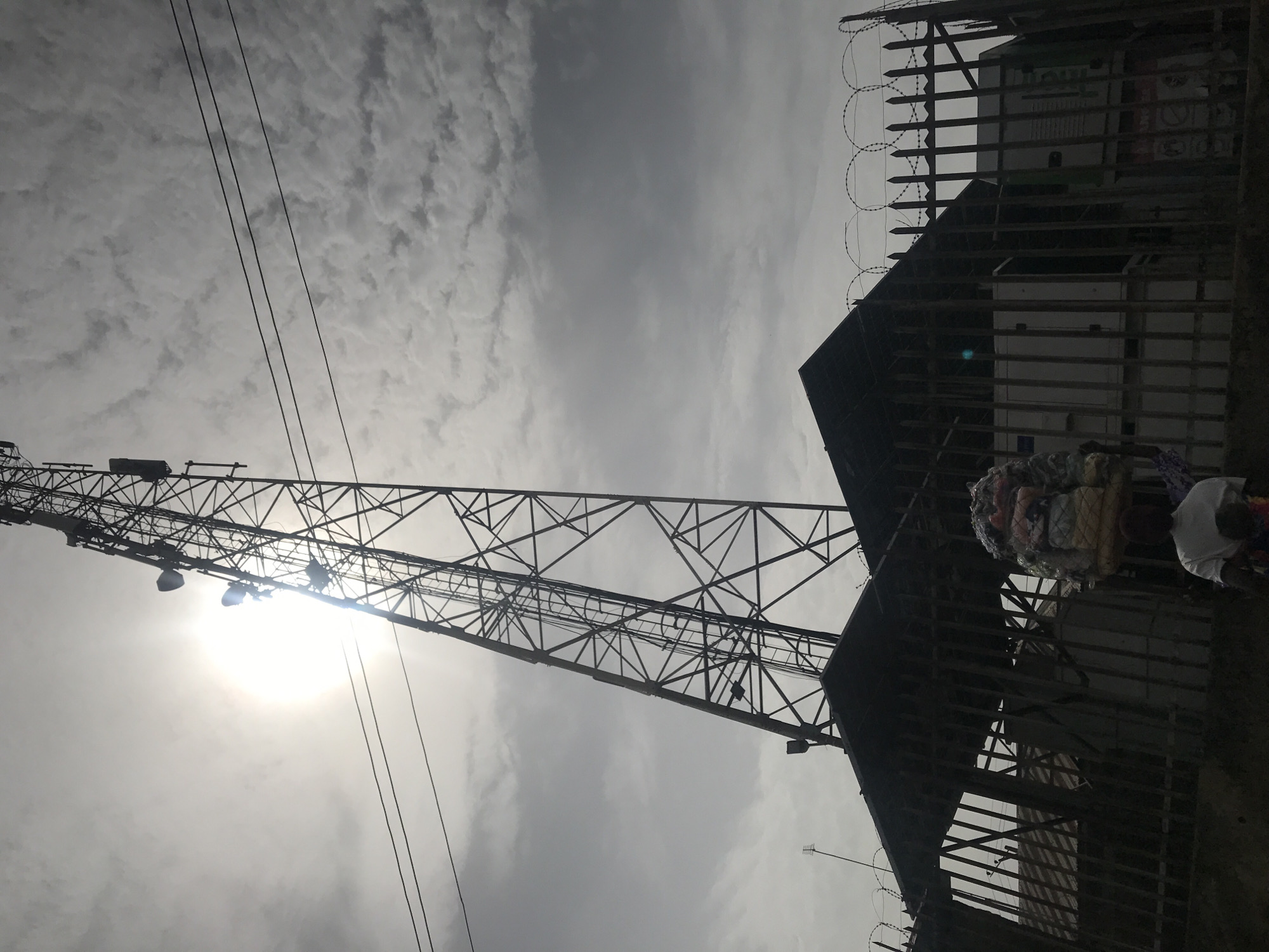 This image, taken from a low angle looking upwards, captures an urban or industrial scene in Ilorin, Nigeria, under an overcast yet bright sky. The dominant feature is a tall, dark metal lattice tower, likely for telecommunications or power transmission, occupying the left side of the frame. Various equipment and antennas are visible on its structure. Several power lines or cables stretch across the upper left sky, seemingly originating or passing near the tower.
On the right side, a dark, silhouetted building or structure is partially visible behind a tall fence. The fence is constructed with vertical bars and is topped with ominous coiled barbed or razor wire. The harsh backlighting from the sun, which appears as a bright glare near the tower in the upper-mid section of the sky, casts most foreground elements into deep shadow, creating a high-contrast, almost monochromatic effect.
Below the fence line on the right, two figures are present. One person is distinctly visible, carrying a large, bundled load of what appears to be textiles or other goods balanced on their head, a common practice in the region. Another person is partially obscured beside them. The sky is largely covered with scattered, diffuse clouds, but the sun's intensity indicates it is daytime, likely mid-day. No legible text is visible on any of the structures or objects. The scene suggests a bustling urban environment where daily life and infrastructure coexist.