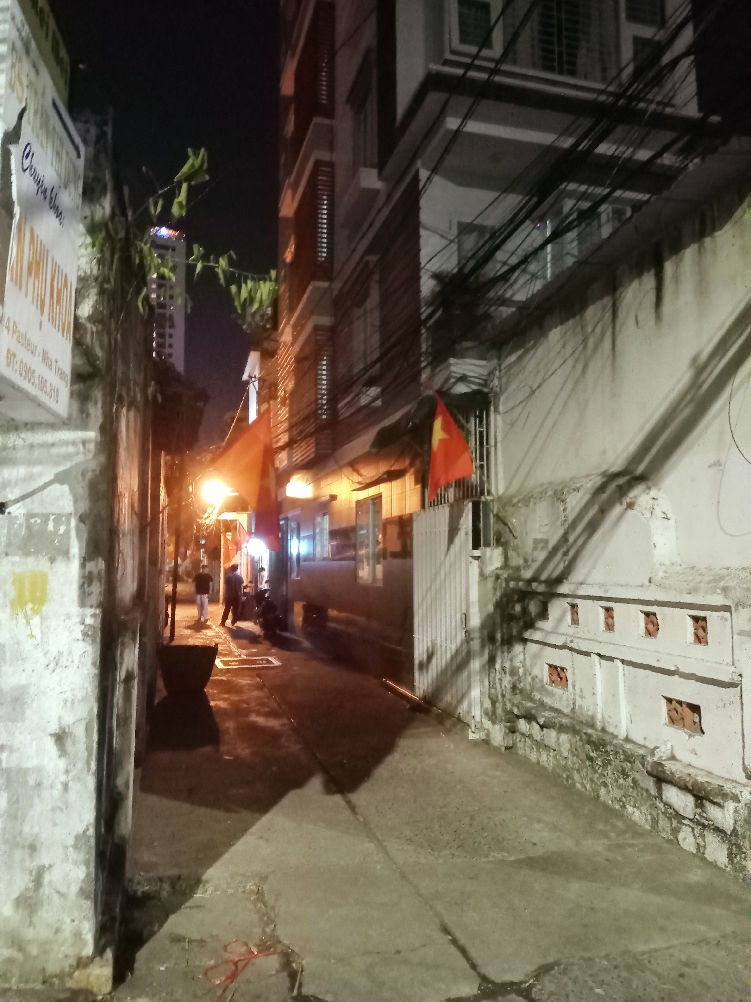 The image depicts a narrow urban alleyway at night in Nha Trang Ward, Vietnam. Artificial lighting, primarily from a bright street lamp positioned in the mid-ground, illuminates the scene and casts long shadows along the pavement and walls.
On the left side of the alley, a light-colored, textured wall features a white sign with Vietnamese text. The text clearly reads "Chuyên khoa" (Specialty), "SẢN PHỤ KHOA" (Obstetrics and Gynecology), followed by an address "4 Pasteur - Nha Trang," and a phone number "ĐT: 0905.105.318." Further along the left, green foliage extends from above, and a taller building's silhouette is visible against the dark night sky.
The right side of the alley is lined by another building, from which a vibrant red Vietnamese flag with a yellow star hangs prominently near a white metal gate. Overhead, a dense network of black electrical wires crisscrosses between the structures. The paved ground appears to be concrete, with some wet patches reflecting the light. In the foreground on the left, a dark, cylindrical bin or pot is visible, and a red string or wire lies on the ground near the bottom left corner.
Two individuals are walking away from the camera in the mid-distance, their figures silhouetted against the light. A scooter is also partially visible nearby. The scene conveys a calm, ordinary nighttime atmosphere in a Vietnamese urban setting.