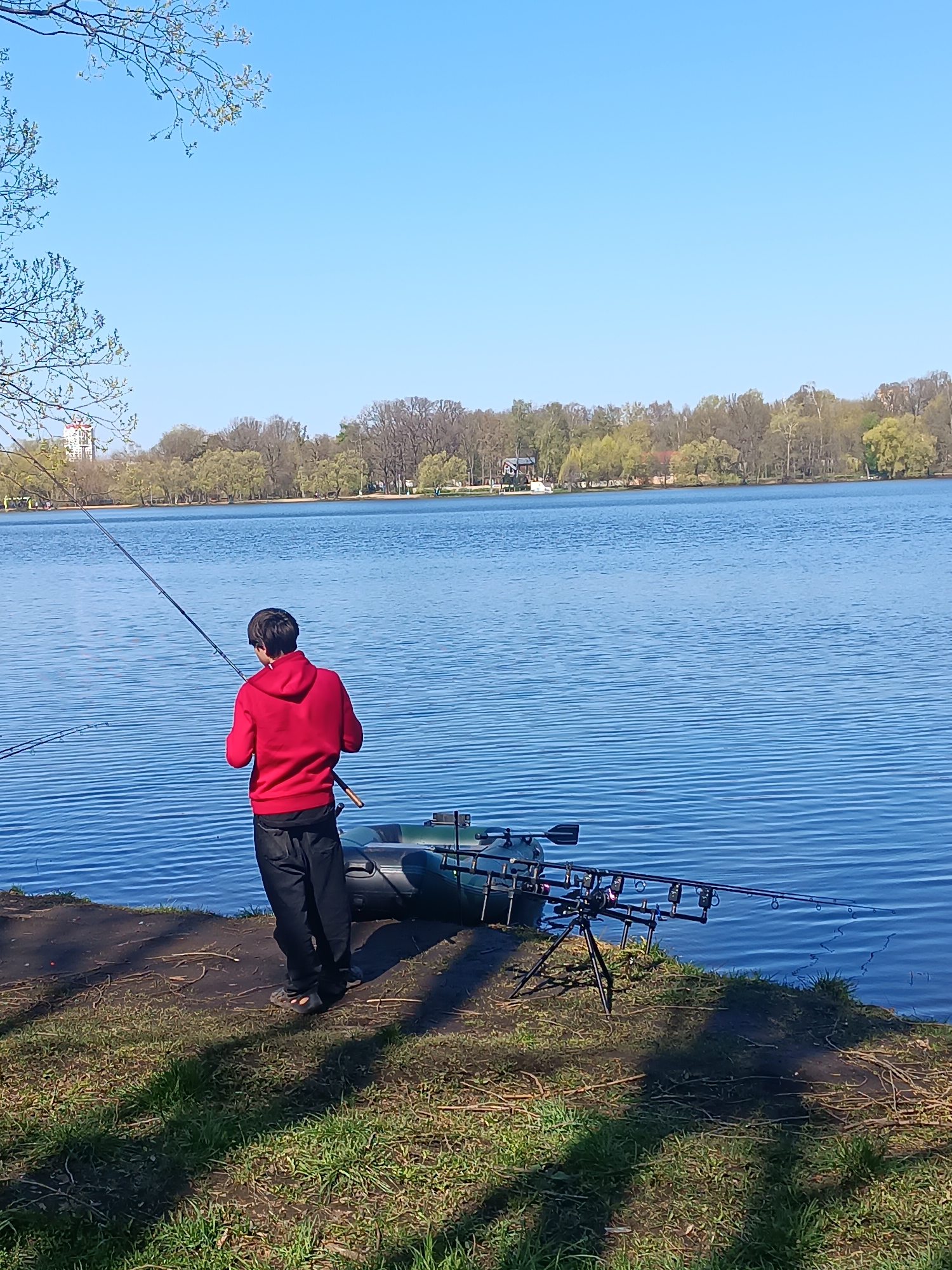 A person in a red hoodie and dark pants is fishing at the edge of a lake. They are standing on a grassy bank next to a small inflatable boat. Two fishing rods are set up on a tripod stand near the boat. The lake is calm with trees visible on the opposite shore. The location is Unknown, Unknown. The sky is clear and blue.