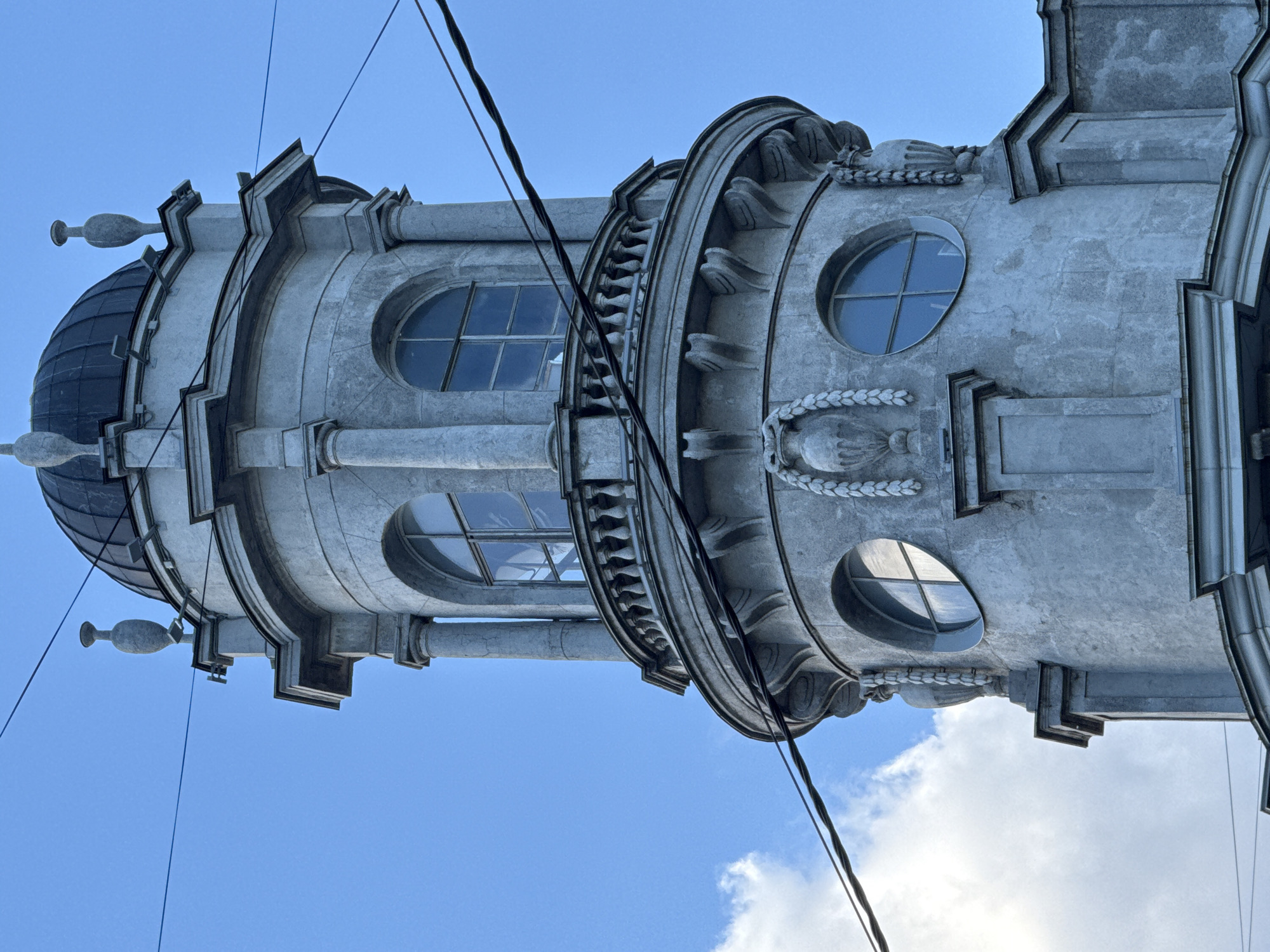 This is a close-up, upward-angled shot of the upper section of a classical-style tower building against a clear blue sky with scattered white clouds. The tower is constructed of light-colored stone and features ornate architectural details. At the very top is a dark, domed roof. Below the dome are decorative urn-like finials on either side of the roof's edge. The main body of the visible tower is cylindrical, with a series of stacked architectural elements.
The uppermost visible section below the dome has arched windows with mullions, framed by pilasters. A balustraded balcony encircles this level. Below the balcony, the tower continues its cylindrical shape, adorned with decorative elements that appear to be stylized floral or laurel swags flanking urn-like sculptures. Two round windows are visible on this level, each with a cross-shaped mullion. The building has a weathered appearance, suggesting age. Several black electrical wires are prominently strung across the frame, diagonally intersecting the view of the tower and the sky. The image captures the upper portion of the building, with the lower parts obscured from view.
This architectural detail is consistent with elements found in Saint Petersburg, Russia. While a definitive identification without further context is impossible, the style is reminiscent of buildings from the late 19th or early 20th century.