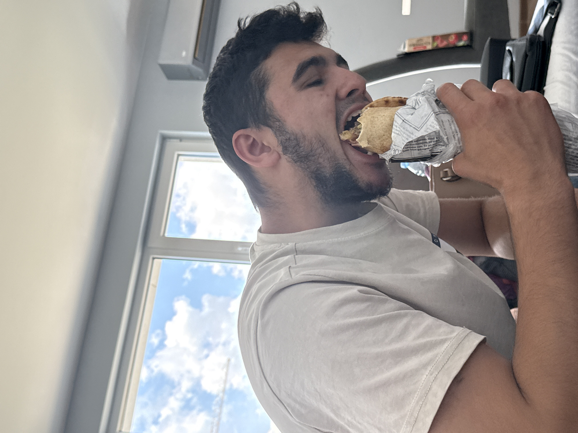 A young man is captured in a candid, low-angle shot, intensely eating a large wrapped food item indoors during the daytime. He has dark, curly hair and a beard, and is wearing a light-colored (possibly off-white or light grey) t-shirt. His mouth is wide open as he takes a significant bite from the wrap, which is partially encased in crinkled paper. His eyes are partially closed, conveying a moment of focused enjoyment of his meal.
The setting appears to be a casual indoor space. A plain white wall occupies the left side of the frame, with a white air conditioning unit mounted high above. To the right, hints of dark furniture or equipment are visible, along with a small box featuring a red label on a surface. A large window provides a view of the outdoors, revealing a bright blue sky dotted with scattered white clouds, indicating clear, pleasant weather.
The food item is a type of wrap, such as a gyro or shawarma, enveloped in paper that bears visible printed text. Upon closer examination, the text on the wrapper appears to be in Greek, with discernible phrases such as "Ελληνική κουζίνα" (Greek cuisine). This detail, combined with the location context of Sofia, Bulgaria, suggests he might be enjoying Greek fast food. The overall impression is a casual, everyday moment of someone deeply engrossed in their meal.
