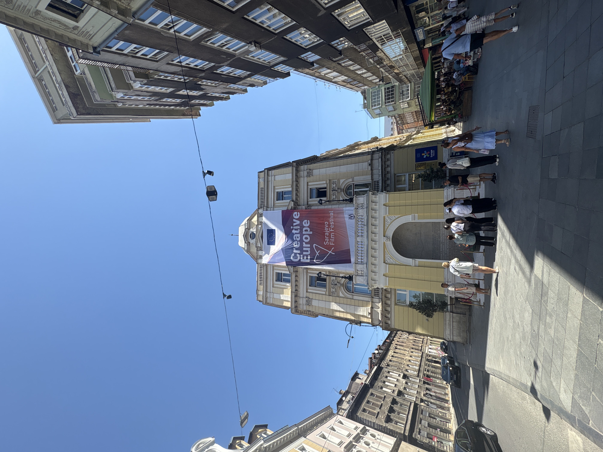 The image depicts a street scene in Sarajevo, Bosnia and Herzegovina. A banner hanging from a building reads "Creative Europe Sarajevo Film Festival." People are gathered in front of the building and along the sidewalk. Some are walking, while others appear to be conversing or observing. Buildings line both sides of the street, with storefronts and cafes visible. Cars are parked and driving on the street. The sky is clear and blue, indicating a sunny day. The architecture suggests a European city, and the banner explicitly identifies Sarajevo.