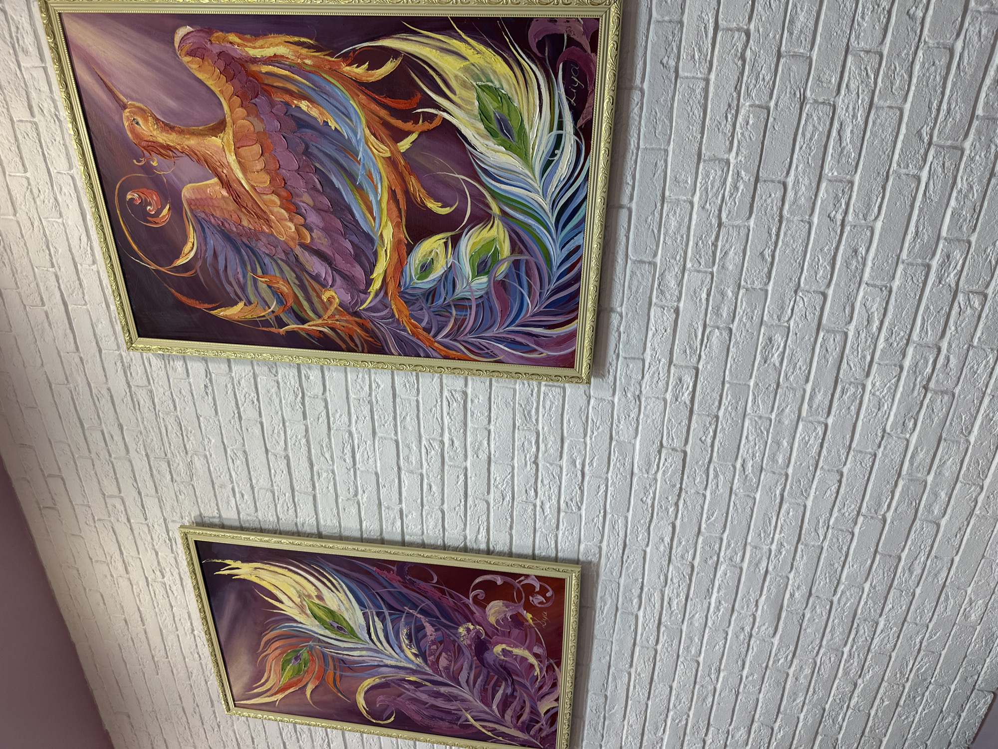 Two framed paintings hang on a white brick wall. The top painting depicts a bird-like figure with vibrant feathers in shades of orange, purple, and yellow. The bottom painting portrays feather-like plumes in similar color tones. Both paintings are adorned with gold-colored frames. The location is in an unknown city.