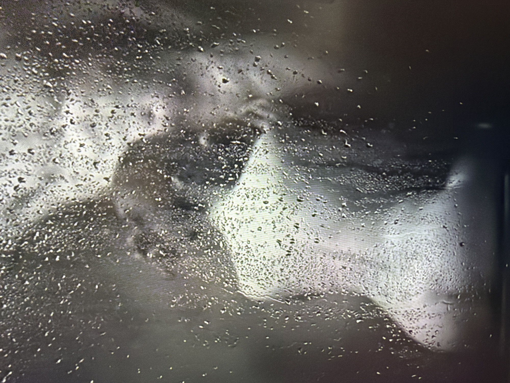 A heavily distorted and pixelated image shows a bare back and shoulders of a person, seen through a surface covered in numerous water droplets. The person appears to be seated and facing away from the camera, with their head tilted downwards. The lighting is dim and diffused, creating a hazy and abstract effect. The background is dark and indistinct. The water droplets on the surface obscure finer details of the subject's skin and form, rendering it impressionistic. The location is unknown, and no specific environmental context can be determined.
