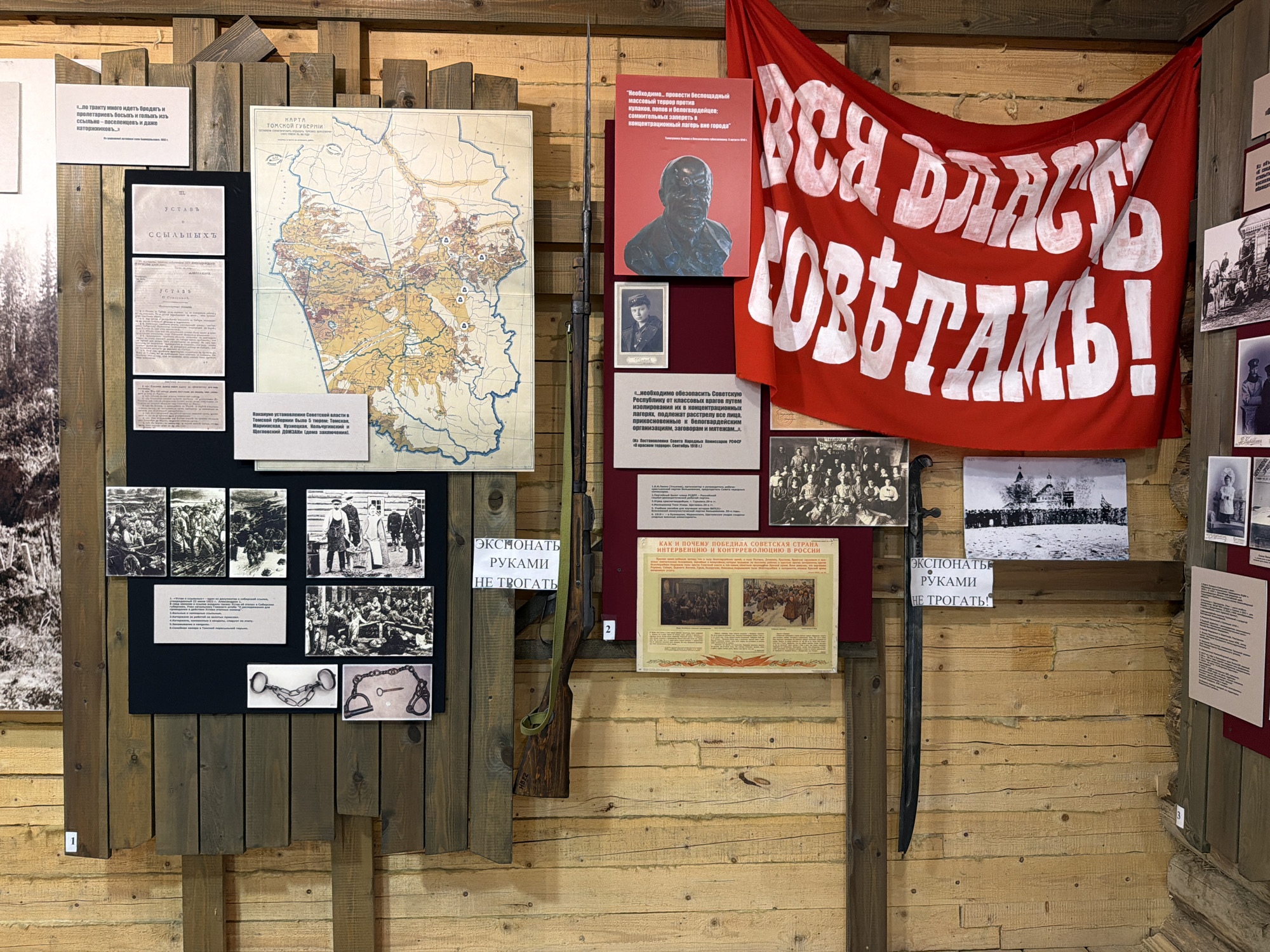 This image shows a museum exhibit in Ust'-Pyzas, Russia, featuring historical artifacts and documents related to the Russian Revolution and Civil War. The exhibit is displayed on a wooden wall, with various items mounted at different heights. In the upper left, a white sign with black text reads "Ц. ... ПО ТРАКТУ МНОГО ИДЕТЬ БРОДЯТЬ И ПРОЛЕТАРНЕВЪ БОСЬКЪ И ПОЛЬЗК ИЗЪ ССЫЛКМЫ - НЕСОГЛАСИЕМЪ И ДАКО ИЗГОТОВЛЕНИЙ." Below this is a black board with three documents. The top document is titled "УСТАВЪ," followed by smaller text. The second document is also titled "УСТАВЪ" with more text. The third document is smaller and appears to be a typed letter. To the right of these documents is a large, faded map titled "КАРТА ТОМСКОЙ ГУБЕРНИИ." The map depicts geographical features and colored regions, likely representing administrative divisions or resources. Below the map is a sign with text in black. Dominating the upper right portion of the image is a large red banner with white Cyrillic lettering. The text appears to be a revolutionary slogan. Below the banner, a dark rectangular plaque displays a bust of a man and a smaller portrait of a person in uniform. A long, wooden rifle, possibly a Mosin-Nagant, leans against the wall vertically in the center of the exhibit, its green strap visible. To the right of the rifle, a dark curved sword is mounted diagonally. Below the rifle and to the left, a black board contains several black and white photographs arranged in a grid. The top row has three images, and the bottom row has one larger image. Below these photos are two metallic objects: a set of handcuffs and a key with a chain. To the right of the rifle and sword, there is another display. At the top is a black and white photograph of a group of people standing in front of a building. Below this are two more photographs. One shows a group of people in a snowy landscape, and the other shows a single person in uniform. Further to the right, a red board has a title in large Cyrillic letters: "КАК И ПОЧЕМУ ПОБЕДИЛА СОВЕТСКАЯ СТРАНА ИНТЕРВЕНЦИЮ И КОНТРРЕВОЛЮЦИЮ В РОССИИ." Below this title are two more black and white images depicting scenes of conflict or gathering. Several smaller white signs with black text are placed throughout the exhibit. One sign, positioned between the rifle and the red board,