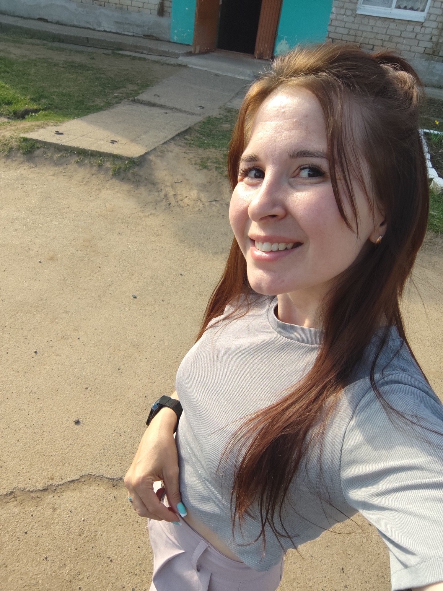 The image presents a woman taking a selfie outdoors. She has light skin, brown hair pulled back in a bun with strands framing her face, and is wearing a light grey ribbed long-sleeved shirt and light pink pants. A black smartwatch is visible on her left wrist. Her nails are painted with a light blue color.
The background consists of a beige gravel or dirt surface, a concrete walkway, a patch of grass, and a building. The building appears to be made of light-colored bricks, and has a doorway with a teal-painted door frame. A window can be seen next to the doorway.
Based on the location provided, the scene is set in Фоки, Russia.