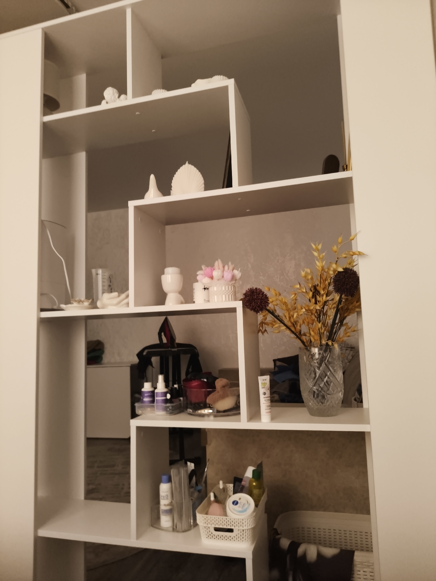 The image shows a tall white shelving unit filled with various decorative items and personal care products. The shelving unit is divided into multiple cubbies and shelves. On the top shelf, there are several white decorative objects, including what appears to be a sculpted figure and shell-shaped candles. The second shelf down has more decorative items, including a white candle holder and a white decorative basket filled with small pink objects. Further down, a clear glass vase contains a bouquet of dried yellow flowers and two dark spherical decorative items. Below this, on another shelf, are two purple spray bottles, a black bag, and a teddy bear. The bottom shelf contains a white basket with various bottles and containers of personal care products, as well as a larger white woven basket to the right. The background appears to be a textured wall. The location is stated as Сеніцкі сельскі Савет, Belarus.