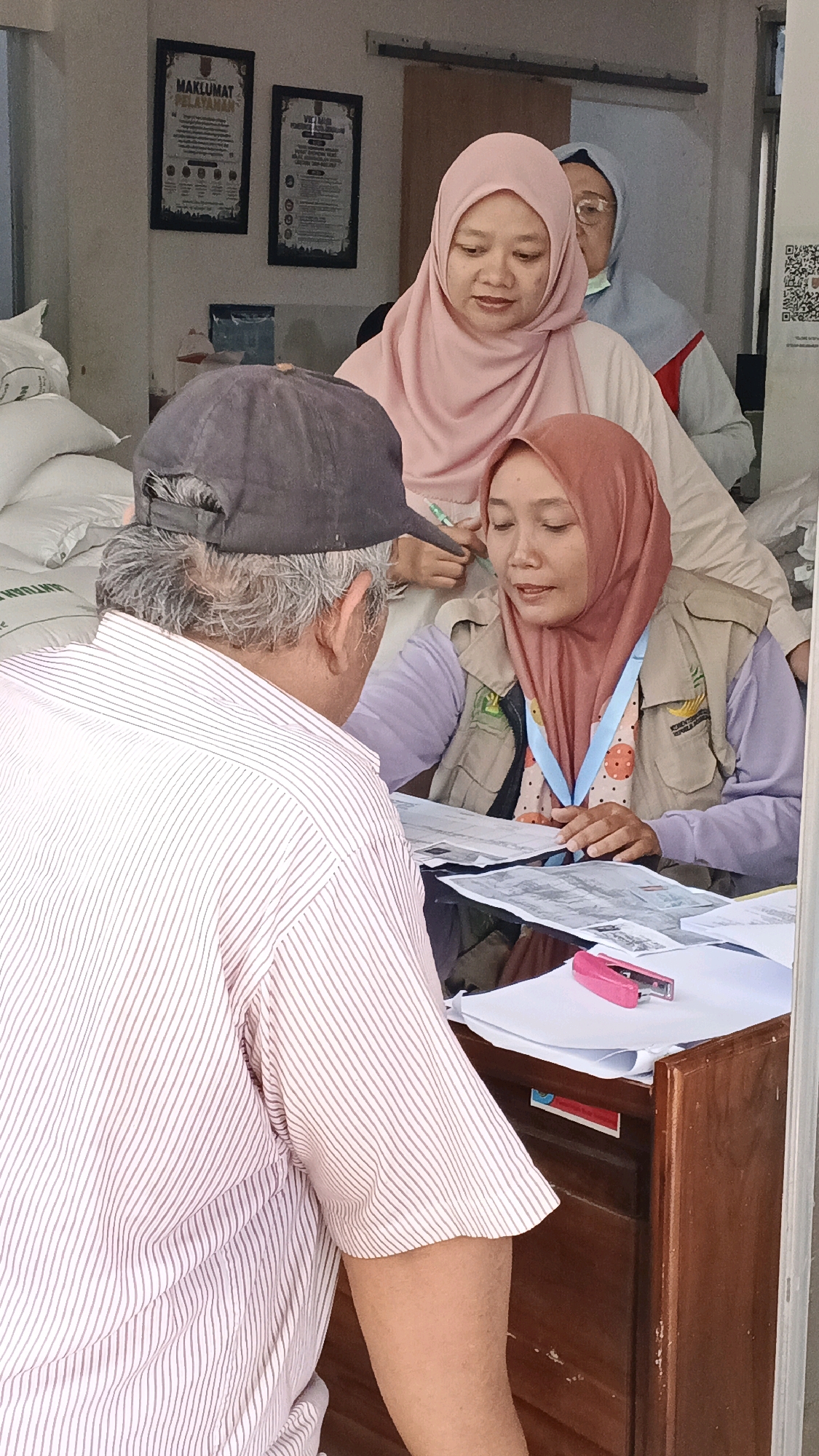 In Semarang, Indonesia, an indoor scene depicts a transaction at a wooden counter. An older man, wearing a striped shirt and a dark baseball cap, is seated, facing away from the camera. He appears to be interacting with two women. One woman, wearing a pink hijab, is seated behind the counter, looking at documents and holding a pen. She is wearing a beige vest over a purple top. To her left and slightly behind her, another woman wearing a light blue hijab is visible, also behind the counter, looking towards the man. A pink stapler rests on the documents on the counter. In the background, there are framed posters on the wall, one with the title "MAKLUMAT PELAYANAN," and several large white bags are stacked to the left. The overall impression is that of a service or administrative counter.