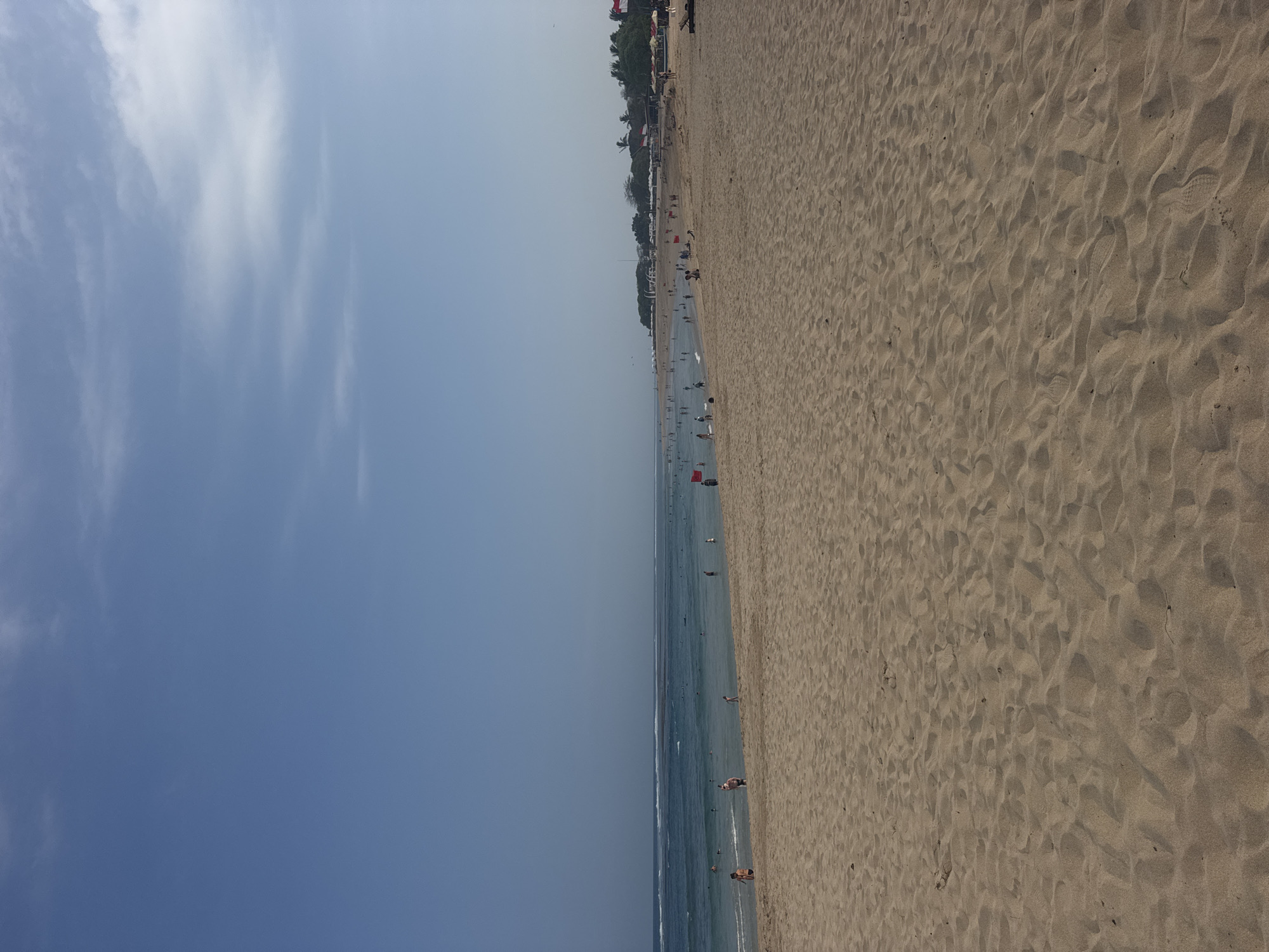 The image depicts a wide, sandy beach with a clear blue sky and gentle waves. Several people are visible in the shallow water and on the wet sand near the shoreline. The beach is populated with many individuals, some of whom are wading in the ocean, while others are walking along the water's edge. Further back, a line of beach umbrellas and what appear to be small structures or buildings are nestled amongst trees. Red flags are placed at intervals along the beach. The sand is a light brown color and shows the texture of small ripples. This scene is consistent with the Kuta Selatan beach area in Indonesia, known for its popular tourist destinations.