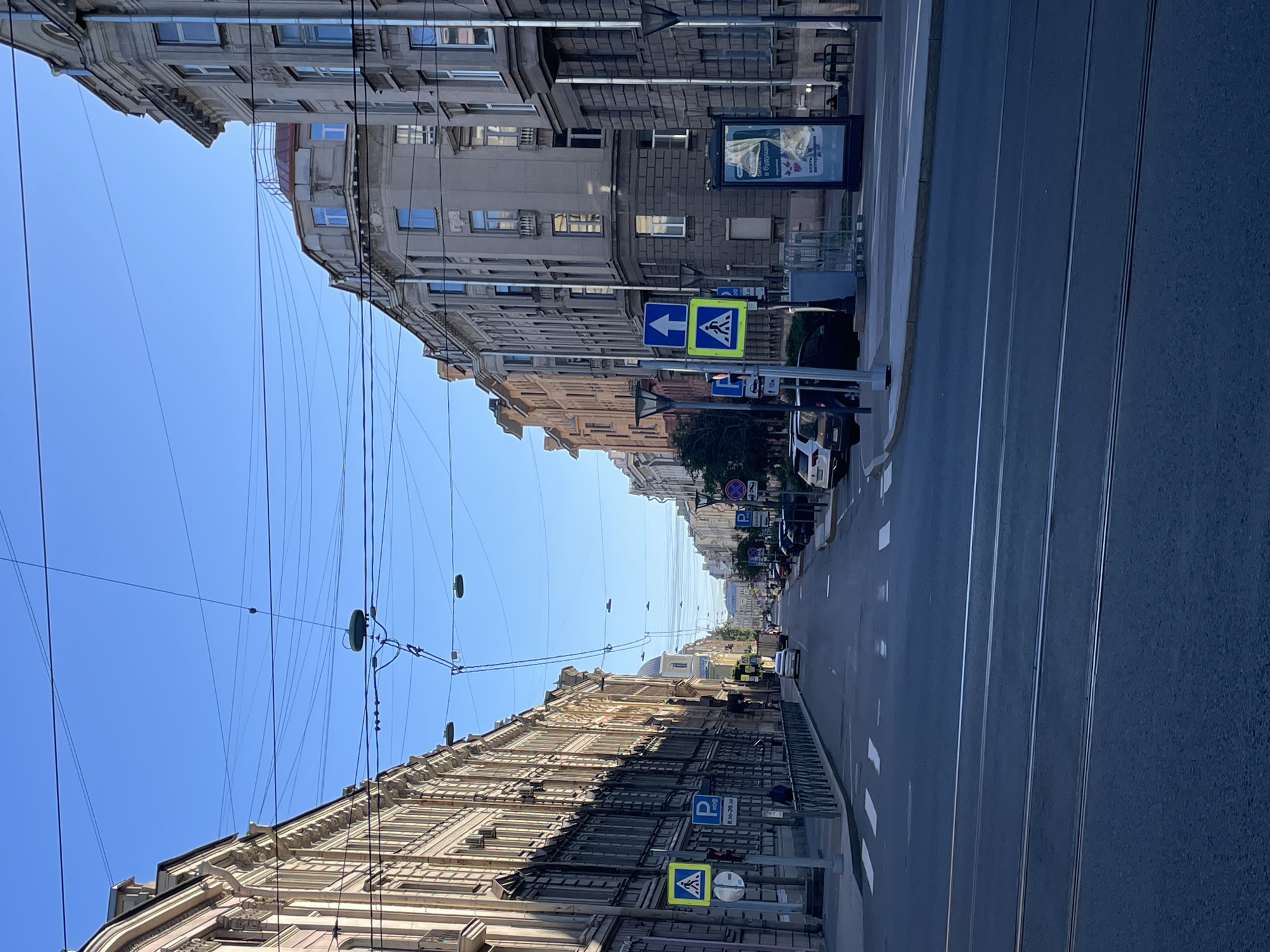 A street scene in Saint Petersburg, Russia, depicts a wide road lined with tall, multi-story buildings on both sides. The architecture suggests a historic urban environment, with classical or neo-classical facades, ornate detailing, and numerous windows. The sky is clear and blue, indicating fair weather. Overhead, a complex network of electrical wires and poles crisscrosses the sky, suggesting the presence of public transportation like trams or trolleybuses.
The street itself is paved with asphalt and features multiple lanes. White road markings are visible, including pedestrian crossings. On the sidewalks, several traffic signs are placed, including pedestrian crossing signs (yellow with a blue pedestrian symbol) and parking signs (blue 'P' symbol with time restrictions). There are also street lamps and trash receptacles.
Several cars are parked along the street and some are driving. A few pedestrian figures are visible on the sidewalks, but they are not the primary focus of the image. One building on the right side of the street has a large advertisement display. The overall impression is a typical urban street in a well-established European city, with a blend of historic buildings and modern infrastructure. The perspective is from street level, looking down the road, which recedes into the distance.