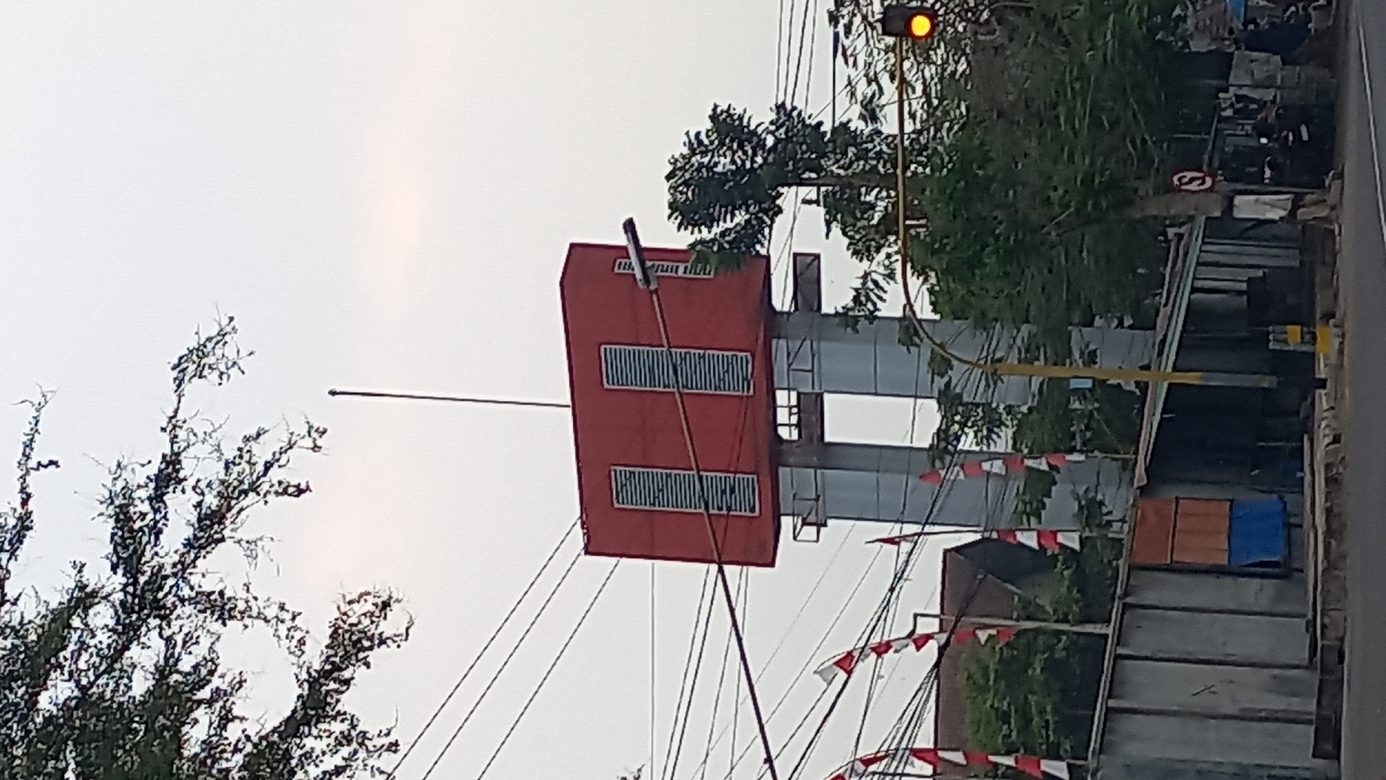 The image shows a street scene in Pare, Indonesia. In the center, a red, rectangular structure sits atop two grey pillars, likely a water tower or a communication tower. The structure has two sets of louvers on its visible face. A traffic light with a yellow bulb illuminated is visible to the right of the tower. Several Indonesian flags, red and white, are planted on poles in the background. The street is lined with various buildings and trees. Overhead, power lines crisscross the sky. A few motorcycles are parked on the right side of the street. The sky is overcast.