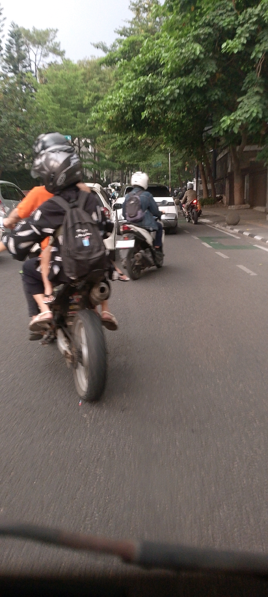 An outdoor, eye-level, medium shot captures a busy street scene in Bandung City, Indonesia, during the day. The focus is on two individuals riding a motorcycle, with the camera positioned slightly behind and to the left of them, giving a rear view. The rider in front is wearing a black shirt with white feather-like patterns and a black helmet. The passenger behind them is a child wearing a white helmet, sitting on the motorcycle with legs straddling the seat, and wearing white sandals. Both are carrying backpacks. The motorcycle is black and appears to be in motion, evidenced by motion blur on the rear wheel and the overall scene.
Other motorcycles and cars are visible on the asphalt road, suggesting moderate traffic. To the right of the road, a sidewalk with a concrete barrier and planters with small trees is present, followed by a building with a dark facade and windows. More lush green trees line the street, their leaves contributing to the overall impression of a vibrant urban environment. The sky is overcast, creating a diffused light across the scene. The image captures a typical moment of daily life on the streets of Bandung, characterized by motor vehicle transportation and the presence of abundant greenery.