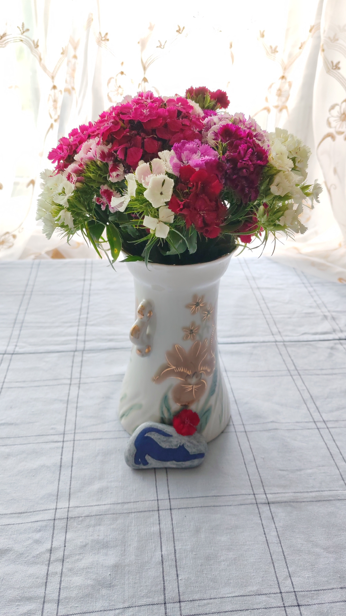 A bouquet of mixed carnations in shades of white, pink, and dark red is arranged in a white ceramic vase. The vase is decorated with gold floral motifs and two small gold figures on its sides. In front of the vase, on a checkered tablecloth, is a small gray stone painted with a blue cat stretching and a red flower. The scene is lit by natural light from a window with sheer curtains.