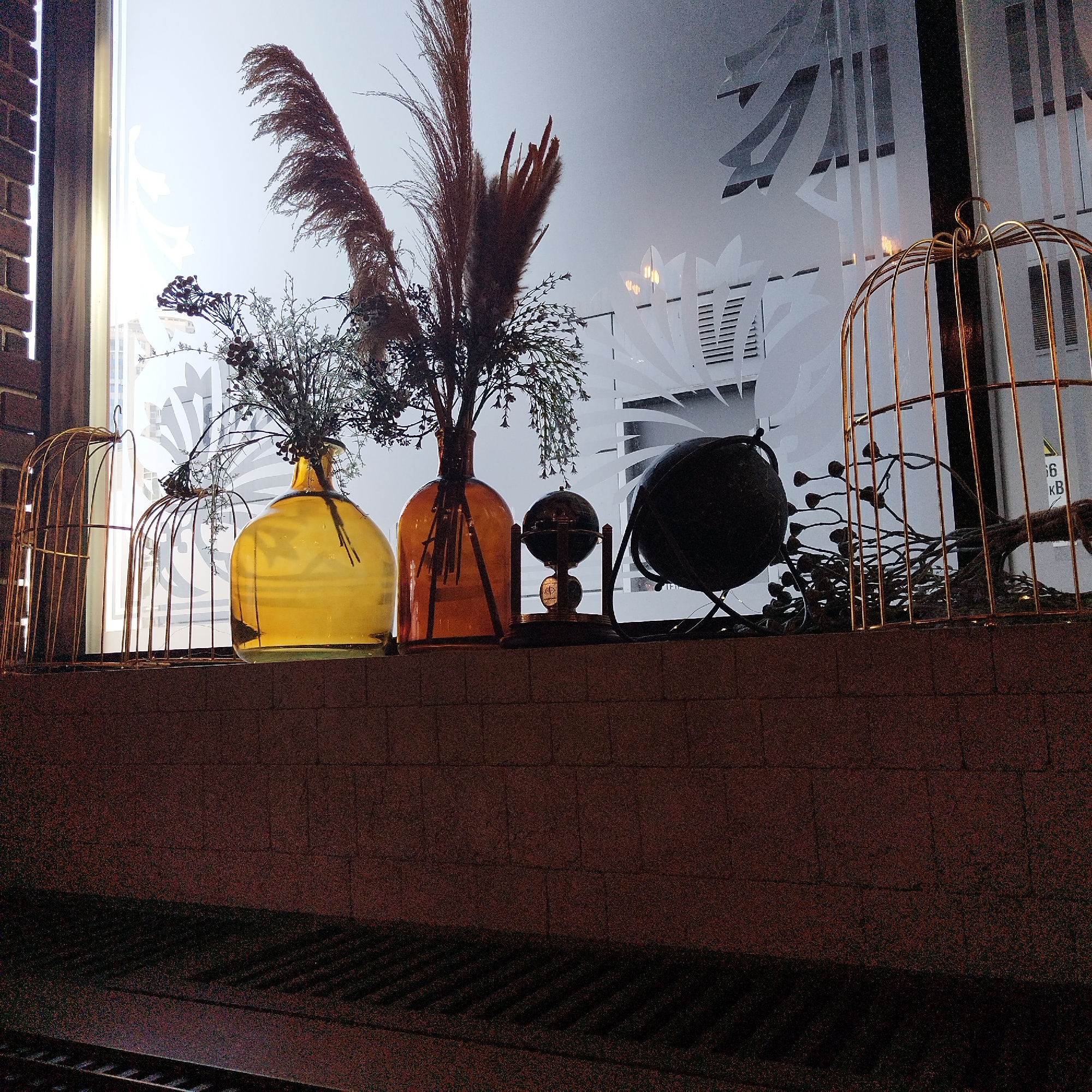 The image displays a decorative arrangement situated on a brick ledge in front of a large window, observed during twilight or nighttime in Yekaterinburg, Russia. The scene appears to be an interior view looking outwards.
The ledge, constructed from dark reddish-brown bricks, supports a collection of vintage-inspired items. From left to right, a golden, open-barred birdcage is positioned next to a large, round yellow glass vase. This vase, along with a slightly taller, amber-colored glass vase to its right, holds tall arrangements of dried botanicals, including prominent feathery plumes resembling pampas grass and other dried foliage and seed pods. Between the vases, a small, dark globe sits on an ornate metallic stand. Further to the right, a larger, dark, spherical object, possibly another globe or decorative sphere, is visible before another golden birdcage, which appears larger than the first and contains additional dried branches and foliage.
The window features frosted glass adorned with intricate, stylized patterns, likely floral or abstract designs, which diffuse the external light and obscure the view outside. The ambient lighting suggests evening or night, with soft, muted light filtering through the frosted glass and warm, artificial light illuminating the display from within.
The foreground below the ledge reveals a dark, textured surface, possibly a ground area or sidewalk, with distinct parallel lines that could be part of a grate or drainage system. No people are visible, and the scene is static. On the far right of the frosted window, faint white text displaying "6 KB" can be seen. The overall impression is one of a carefully curated, rustic or autumnal aesthetic.