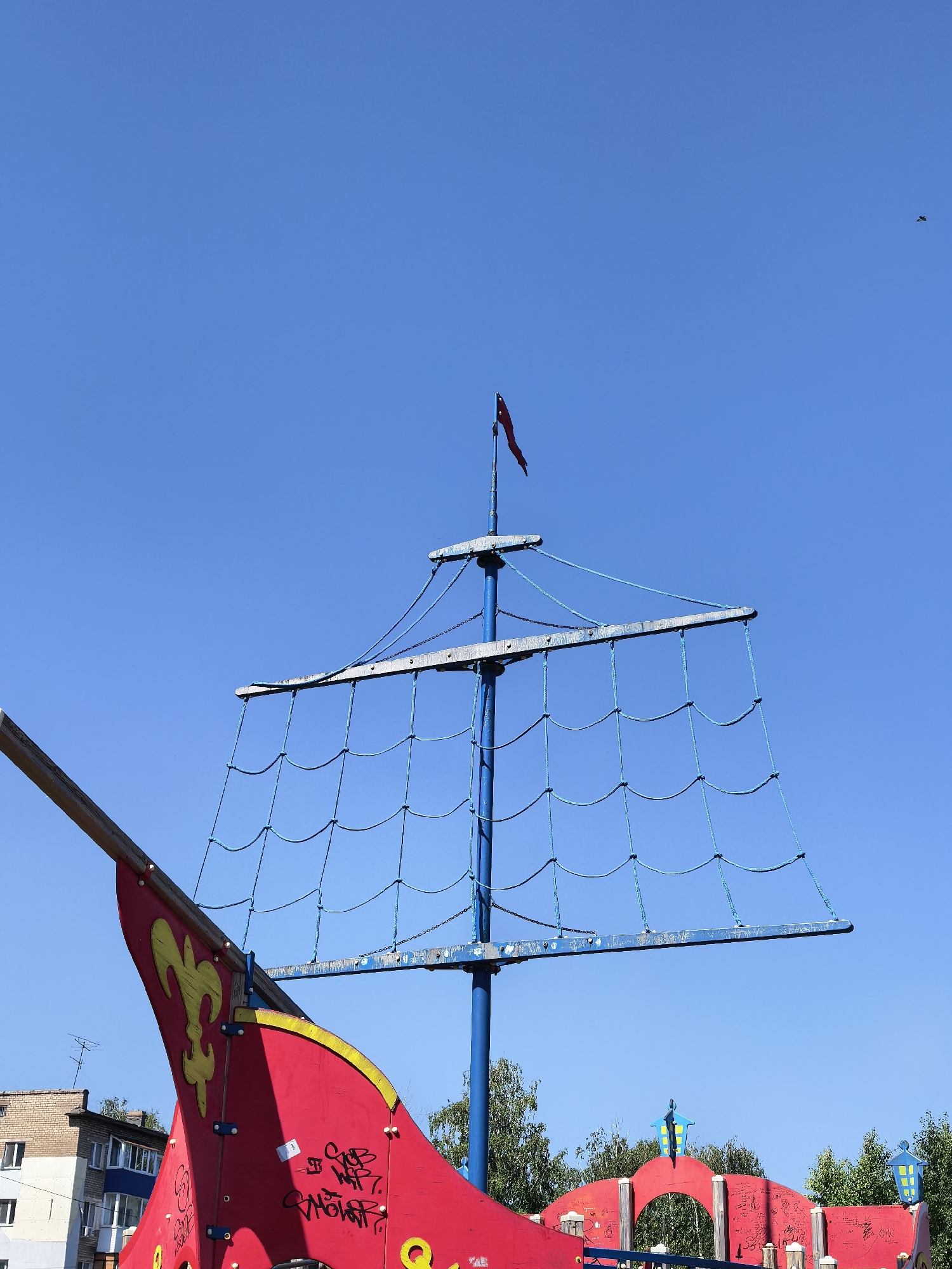 The image captures a brightly painted red playground structure, designed to resemble a pirate ship, under a clear blue sky in the city of Sterlitamak, Russia. The ship's mast, painted blue, is prominently featured with a network of blue ropes forming a net. A small, dark red flag flies from the top of the mast. The bow of the ship is visible on the left, adorned with a yellow fleur-de-lis design. The structure appears to be made of painted wood and metal. Graffiti is visible on the red surfaces of the ship. In the background, the upper floors of a multi-story apartment building and green trees can be seen. A single bird is visible as a small dark speck in the sky on the right side of the image. The overall impression is a sunny day on a playground.