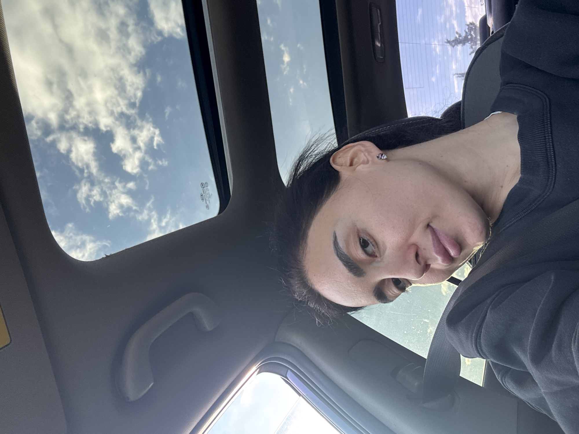 A young adult woman with dark hair is inside a car. She is wearing a dark hoodie and has a seatbelt fastened across her chest. She sticks her tongue out slightly towards the camera. The sky with clouds is visible through the car's sunroof. The location is impossible to determine from the image alone.