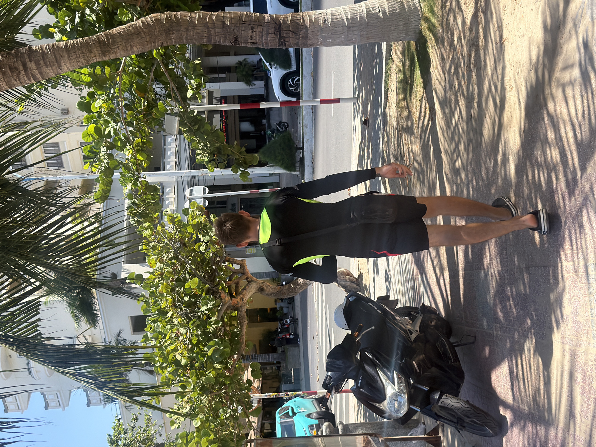 In Nha Trang, Vietnam, a young man, viewed from behind, walks on a tiled sidewalk next to a street. He wears a black long-sleeved top with bright green accents and black shorts. A black crossbody bag is slung over his shoulder. He has short brown hair and is wearing black slip-on shoes. To his left, a black motorcycle is parked on the sidewalk, with a white helmet on its seat. Behind the motorcycle is a turquoise car. Across the street, there are buildings, trees, and other vehicles, including a white car and a scooter with a rider. Palm trees and other foliage line the sidewalk and street. The scene is illuminated by bright sunlight, casting distinct shadows from the palm trees onto the ground.