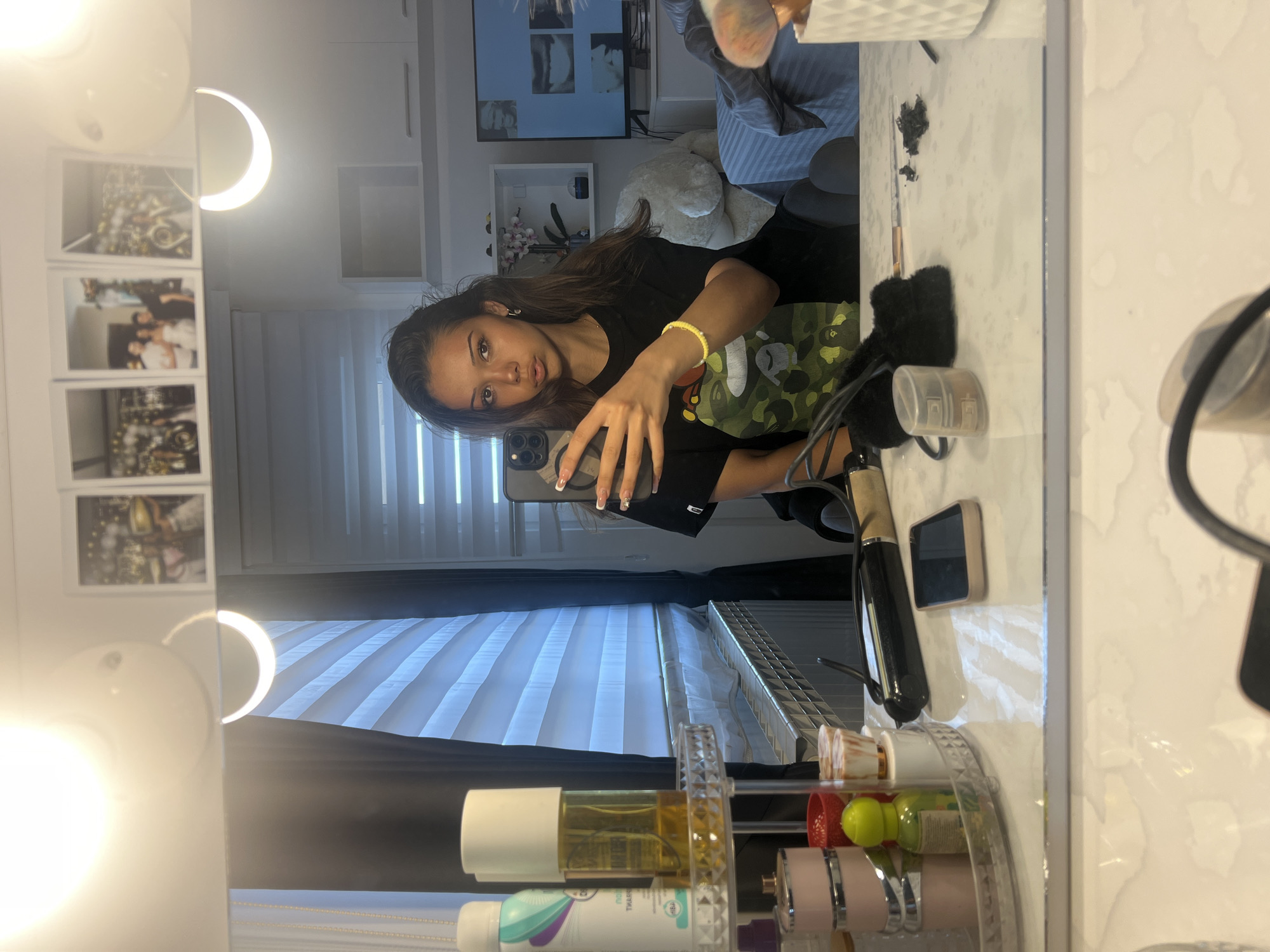 A person takes a selfie in a mirror. The individual, with long brown hair and brown eyes, is holding a phone with their right hand, displaying a manicure. They are wearing a black t-shirt with a green and yellow pattern, and a yellow bracelet. The background shows a room with a window covered by blinds, a white cabinet, framed pictures, and a television. The mirror reflects the person, makeup brushes, hair styling tools, and a multi-tiered shelf with various beauty and hygiene products.