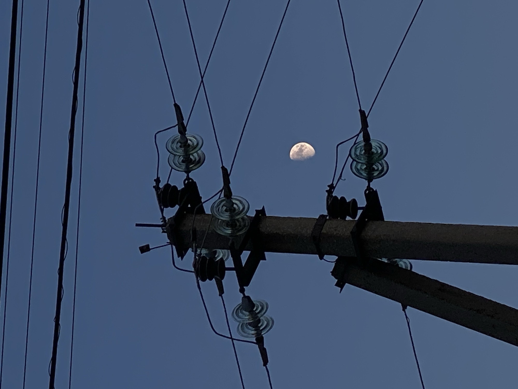 A photograph taken in Cairaclia, Moldova, captures a concrete utility pole supporting multiple electrical wires against a deep blue sky. The moon, appearing as a crescent, is visible in the upper right quadrant of the frame. The utility pole is equipped with several glass insulators, some of which are stacked in pairs, designed to hold and isolate the electrical conductors. The wires are strung across the sky, creating a network of dark lines that intersect with each other and the pole. The lighting suggests it is either dusk or dawn, with the sky exhibiting a twilight hue. The image has a slightly grainy texture, indicative of low-light photography.