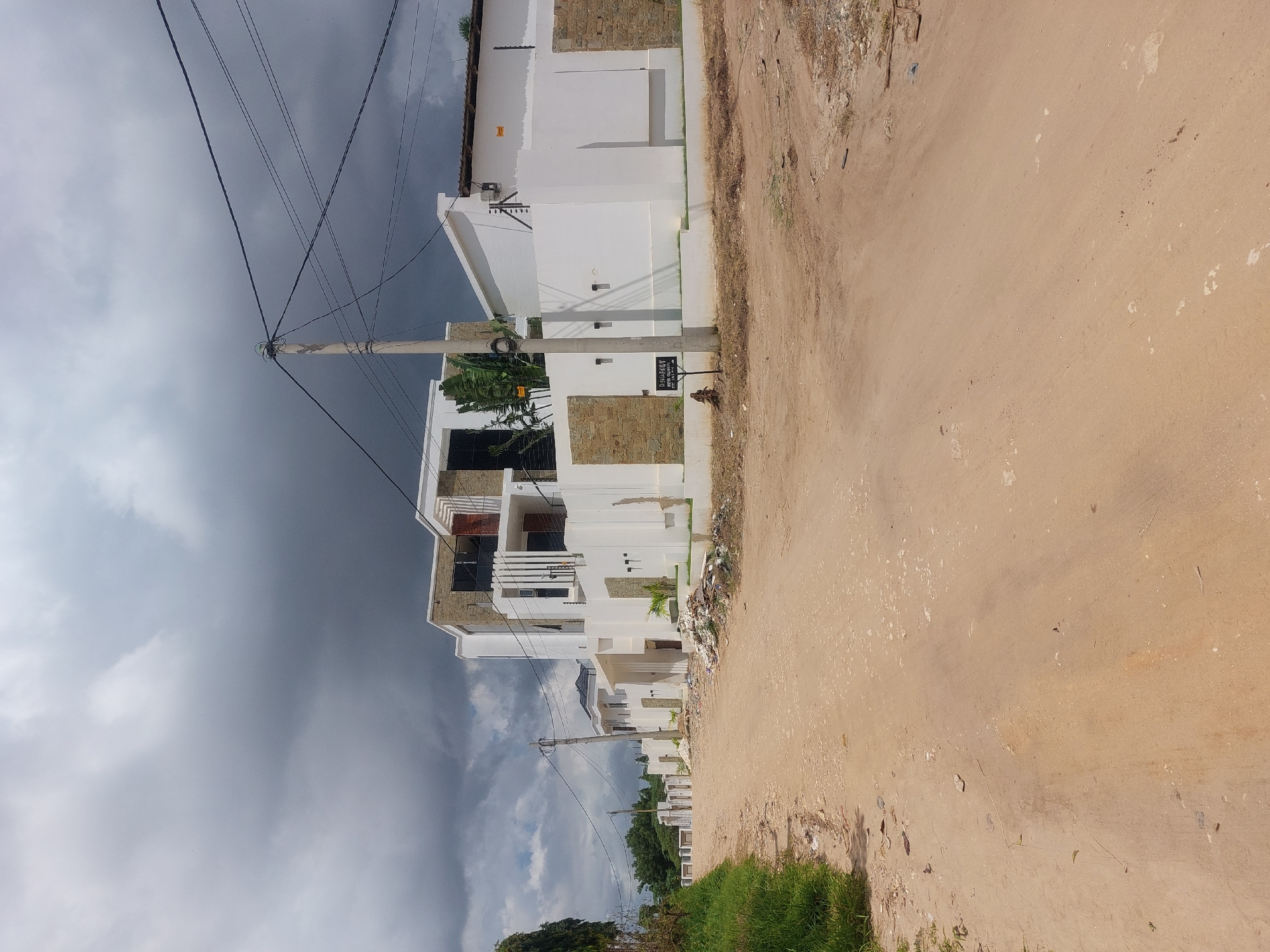 The image shows a residential street in Dar es-Salaam, Tanzania. The houses are modern, two-story structures with white walls and stone accents. A dirt road runs along the side of the houses. The sky is overcast, with dark clouds overhead. There are utility poles with power lines running along the street. Some grass and foliage is visible near the bottom of the frame. No people are visible in the image.