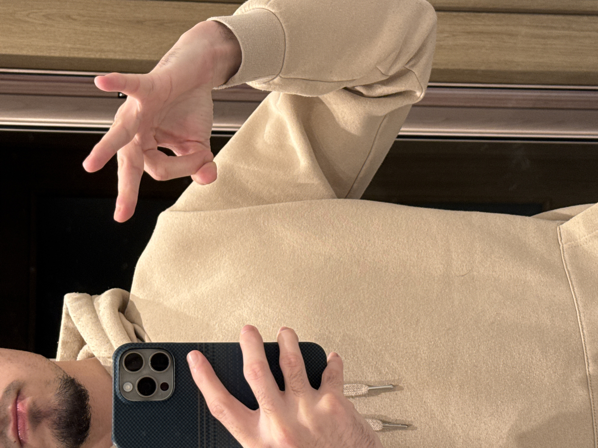 The image is a selfie taken in a mirrored environment. A person is holding a dark blue smartphone with a triple-lens camera system. The reflection shows a portion of the person's face, including their lips, chin, and stubble beard. They are wearing a beige hooded sweatshirt. The person's other hand is held up in a gesture, possibly forming the letter "L" or making a claw-like shape. The background consists of a wooden panel above a reflective surface, likely a mirror. The setting is indoors. There is no evidence to indicate this photo was taken in Sofia, Bulgaria.